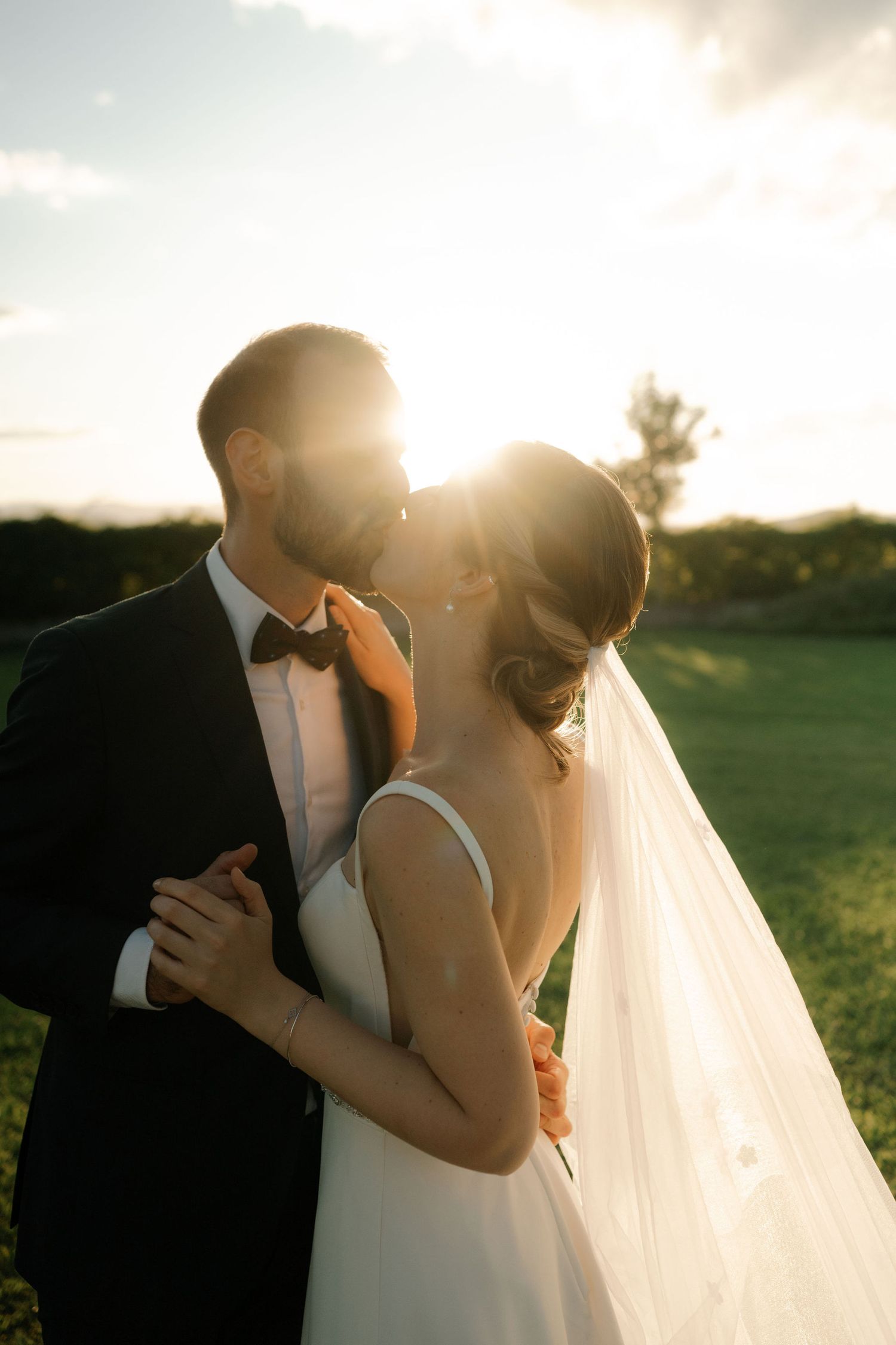 Silhouetted newlyweds embrace in golden sunlight during sunset outdoor wedding photoshoot.