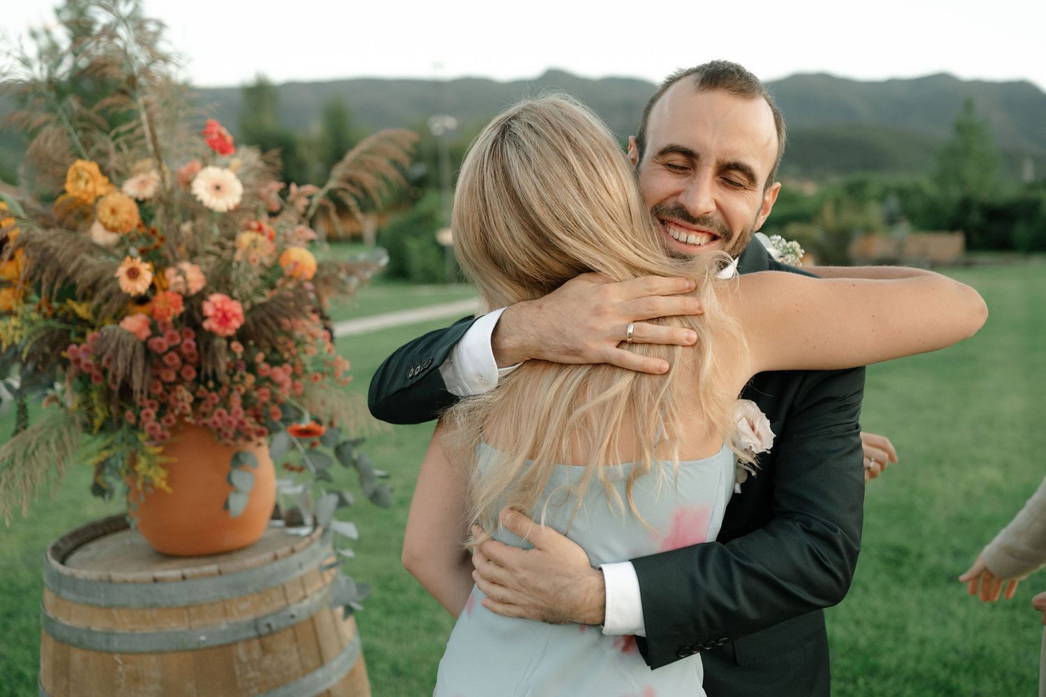 A couple embraces at an outdoor wedding with a rustic barrel and orange floral arrangement in the background.