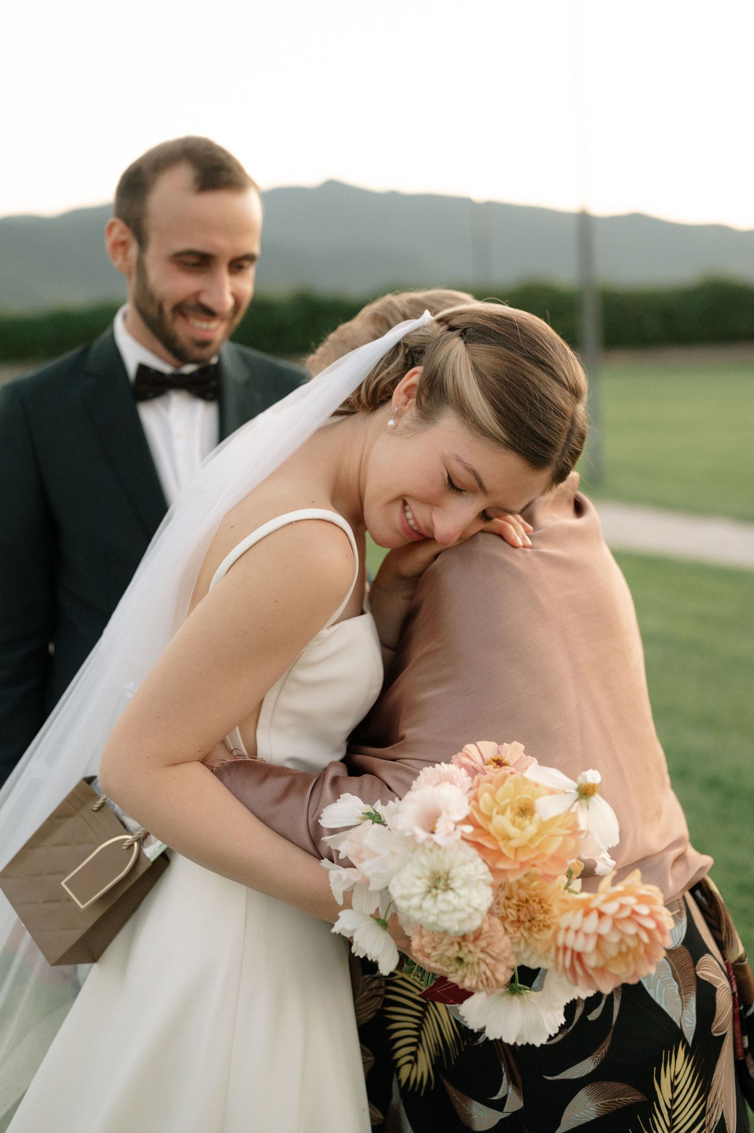 A couple in wedding attire share an intimate moment outdoors with a bouquet of pink and white flowers.
