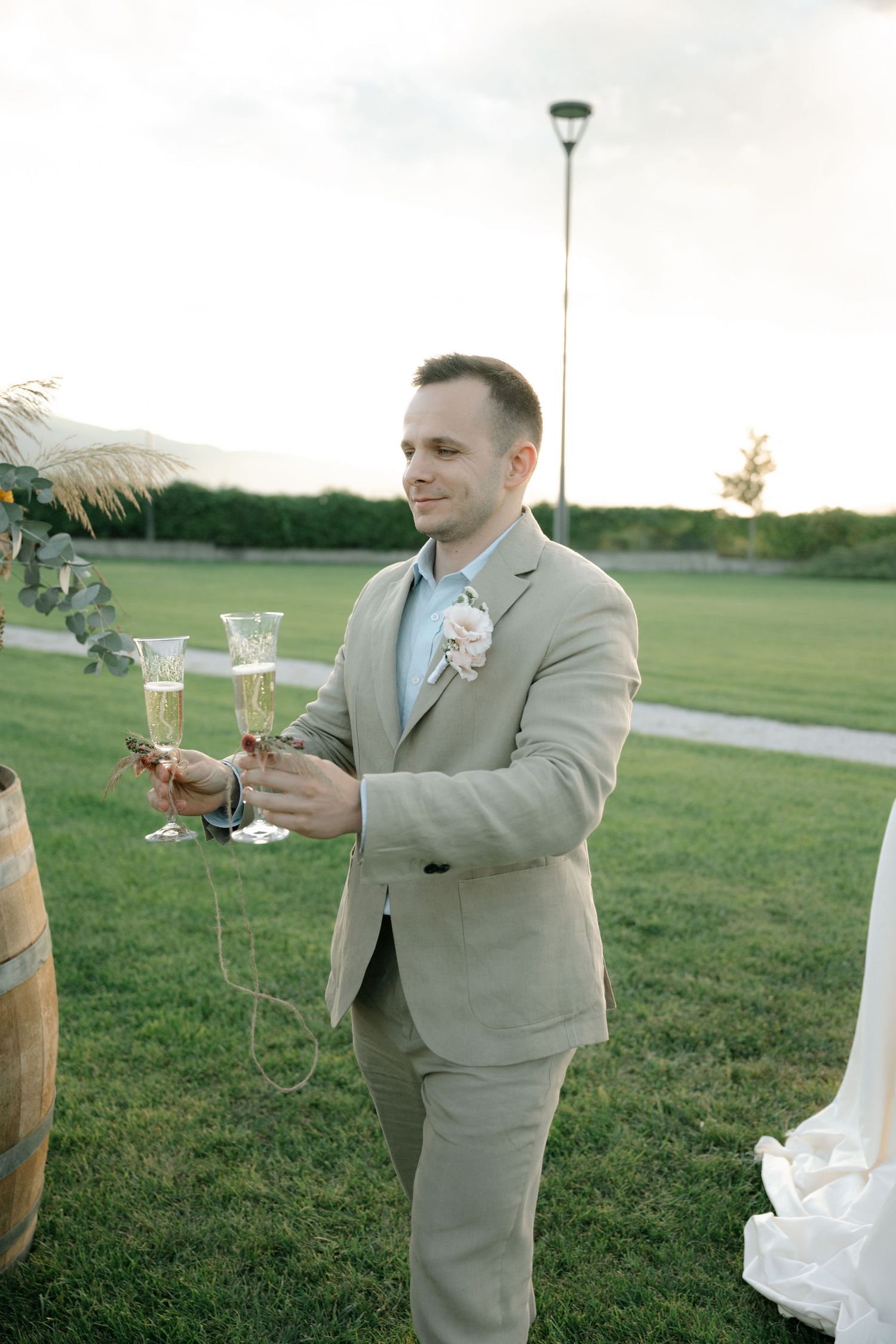 A couple in wedding attire toasts with champagne glasses on a green lawn during sunset.