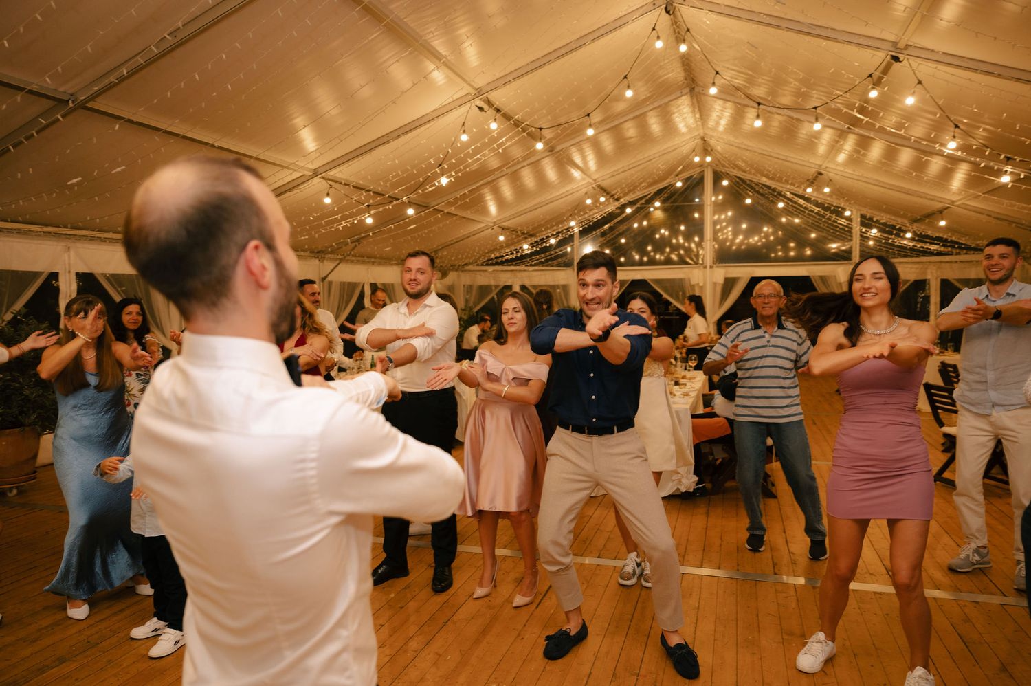 People dancing in a line formation under string lights at an indoor reception venue.