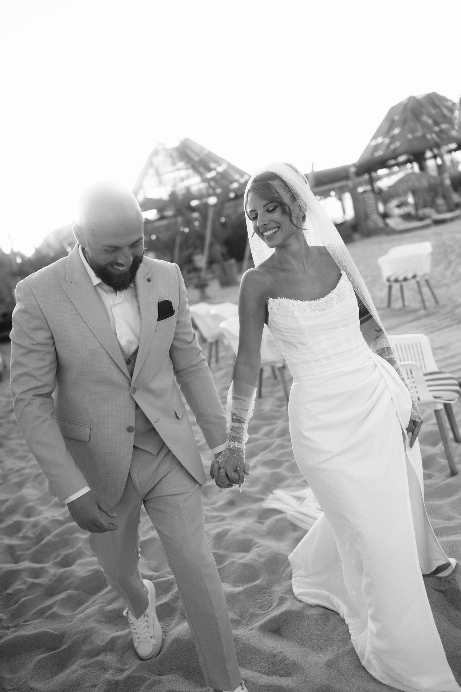 A couple in formal wedding attire playfully dances and poses together on a sandy beach.