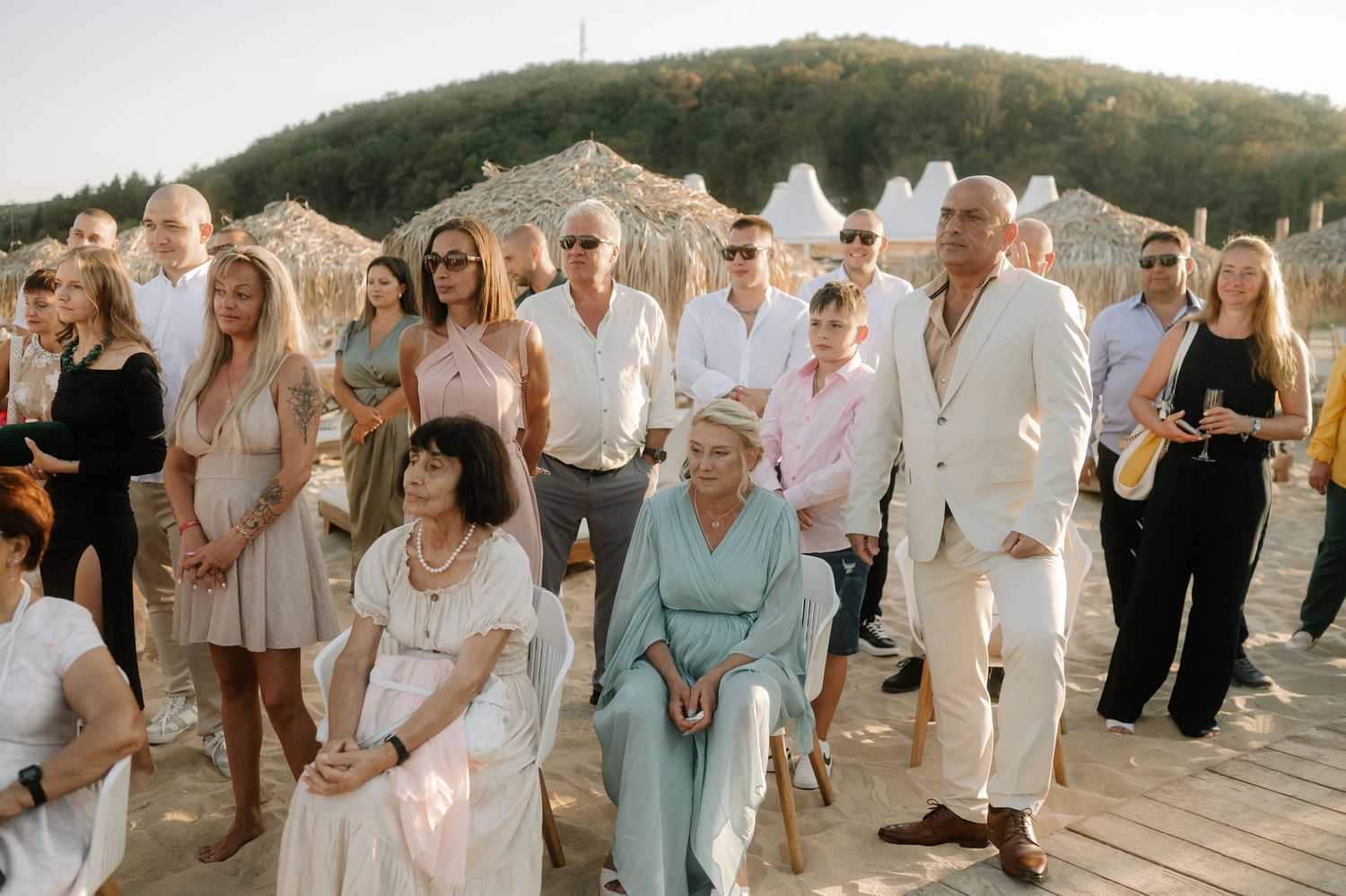 Group of wedding guests in light colored attire gather outdoors during sunset celebration at scenic venue.