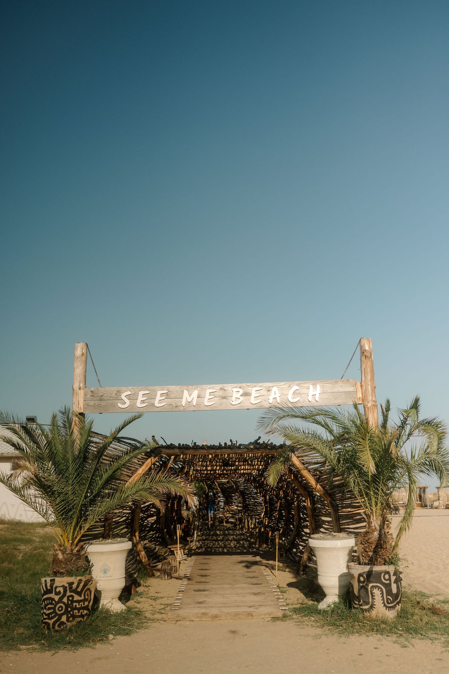 A rustic wooden entrance sign reading 'SEE ME BEACH' stands among palm trees in a desert-like beach setting.
