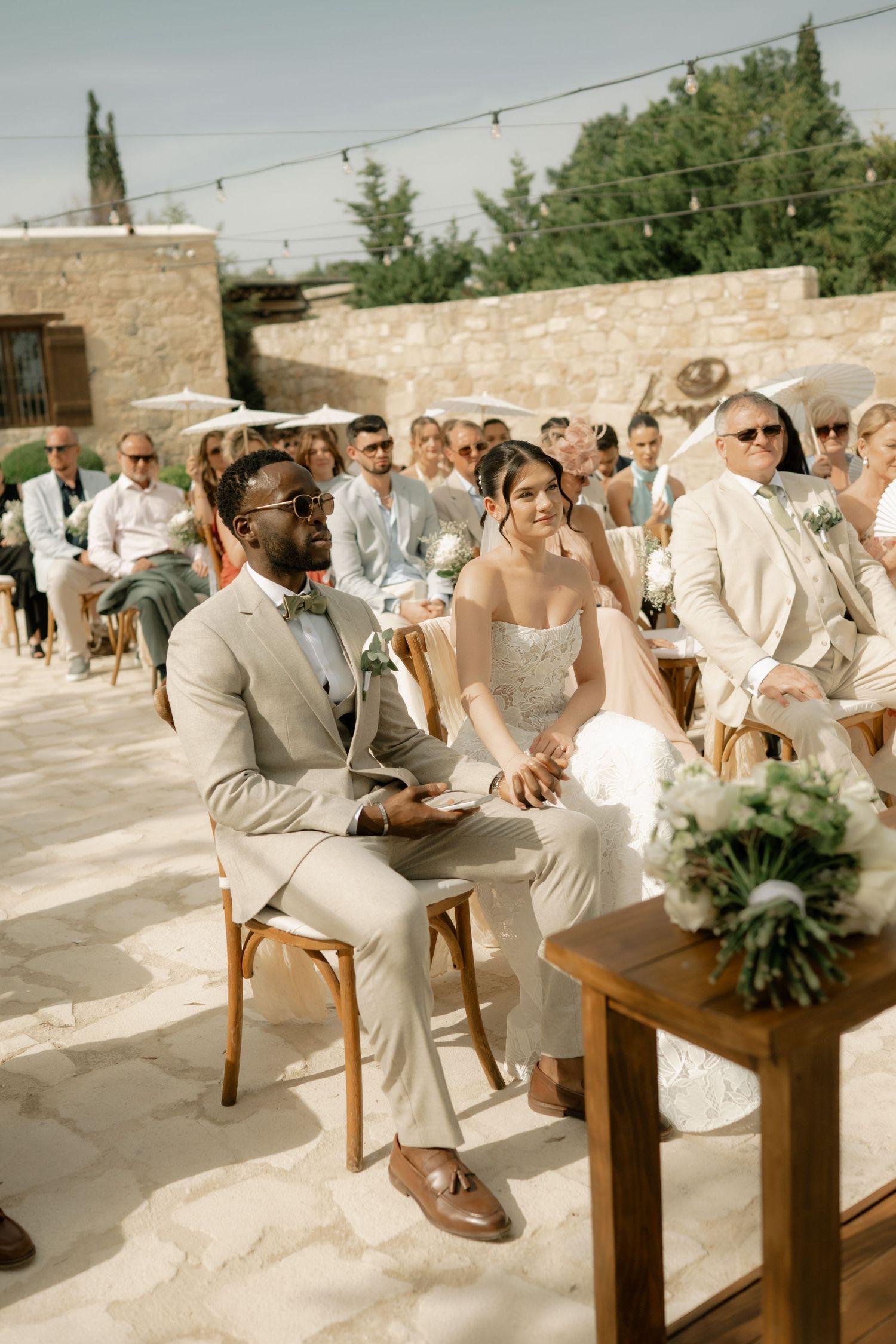 Bride and groom sit together during outdoor wedding ceremony, guests seated behind them in elegant stone venue.