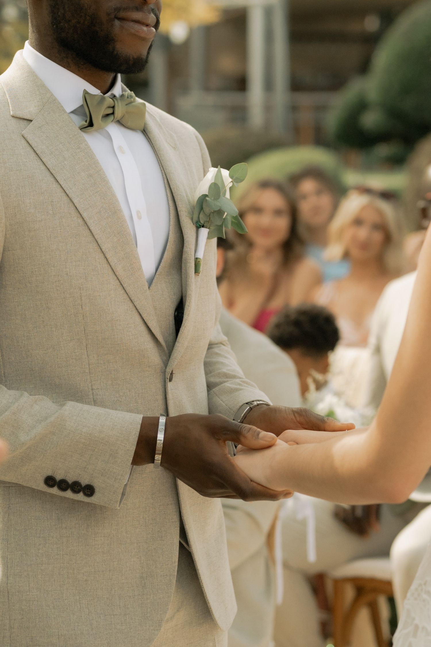 Groom in beige linen suit holding bride's hand during outdoor wedding ceremony with guests in background.