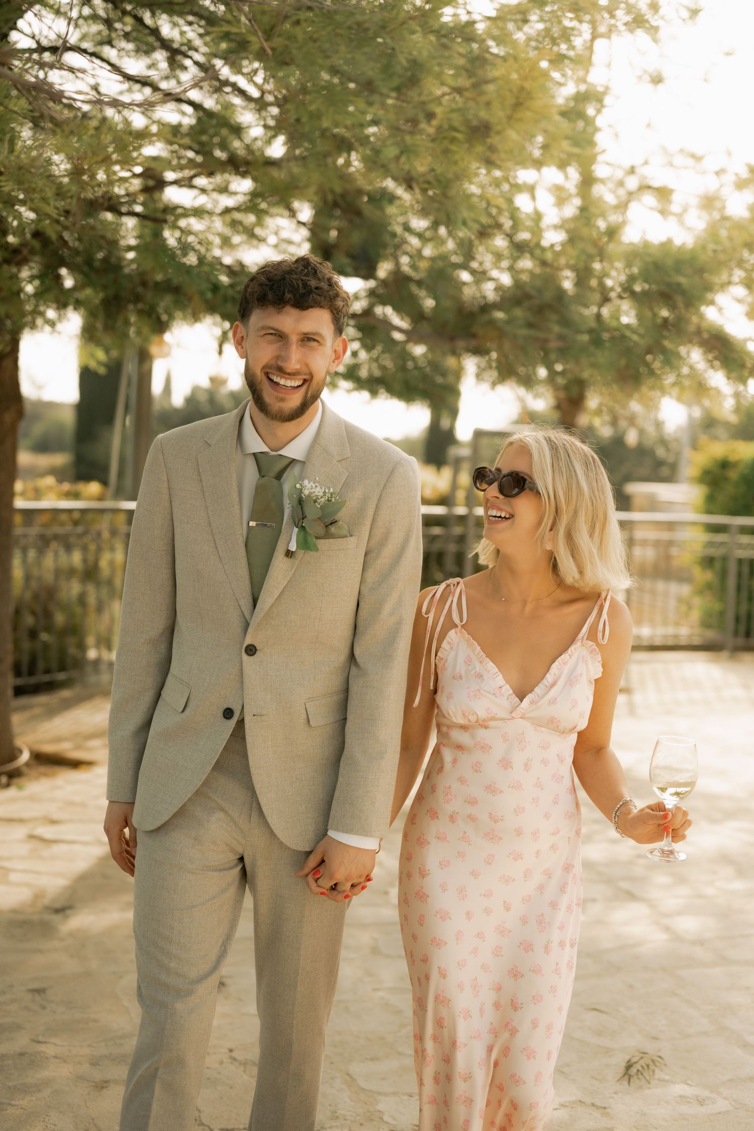 Smiling couple in wedding attire walking outdoors, man in tan suit with boutonniere, woman in floral dress holding wine glass.