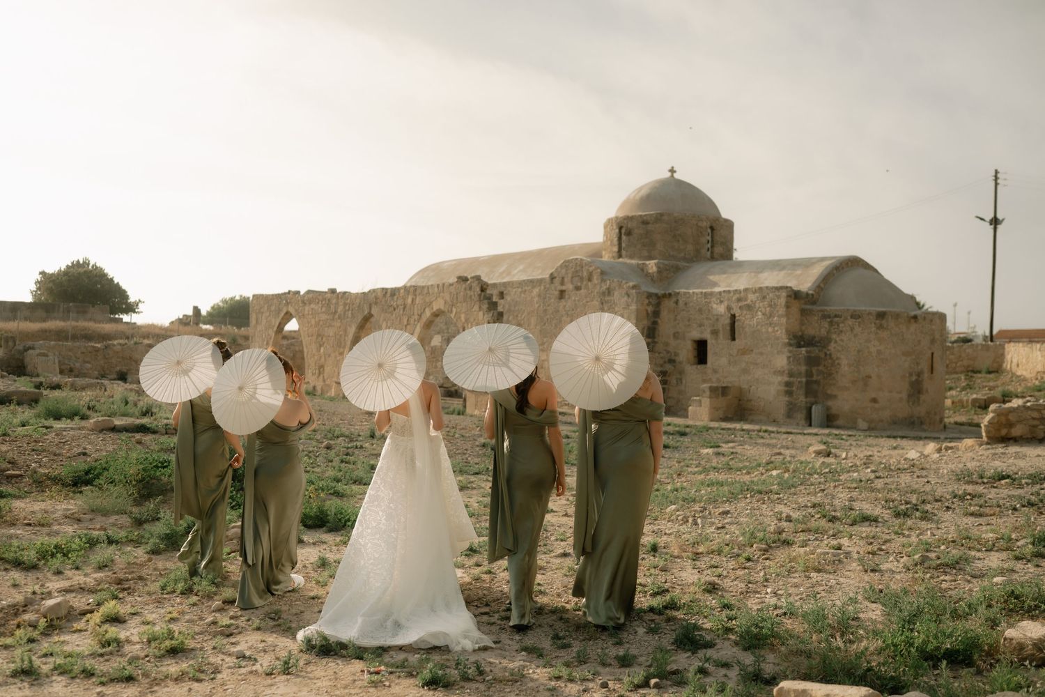Bride and bridesmaids in sage green dresses holding white parasols near ancient stone church ruins.