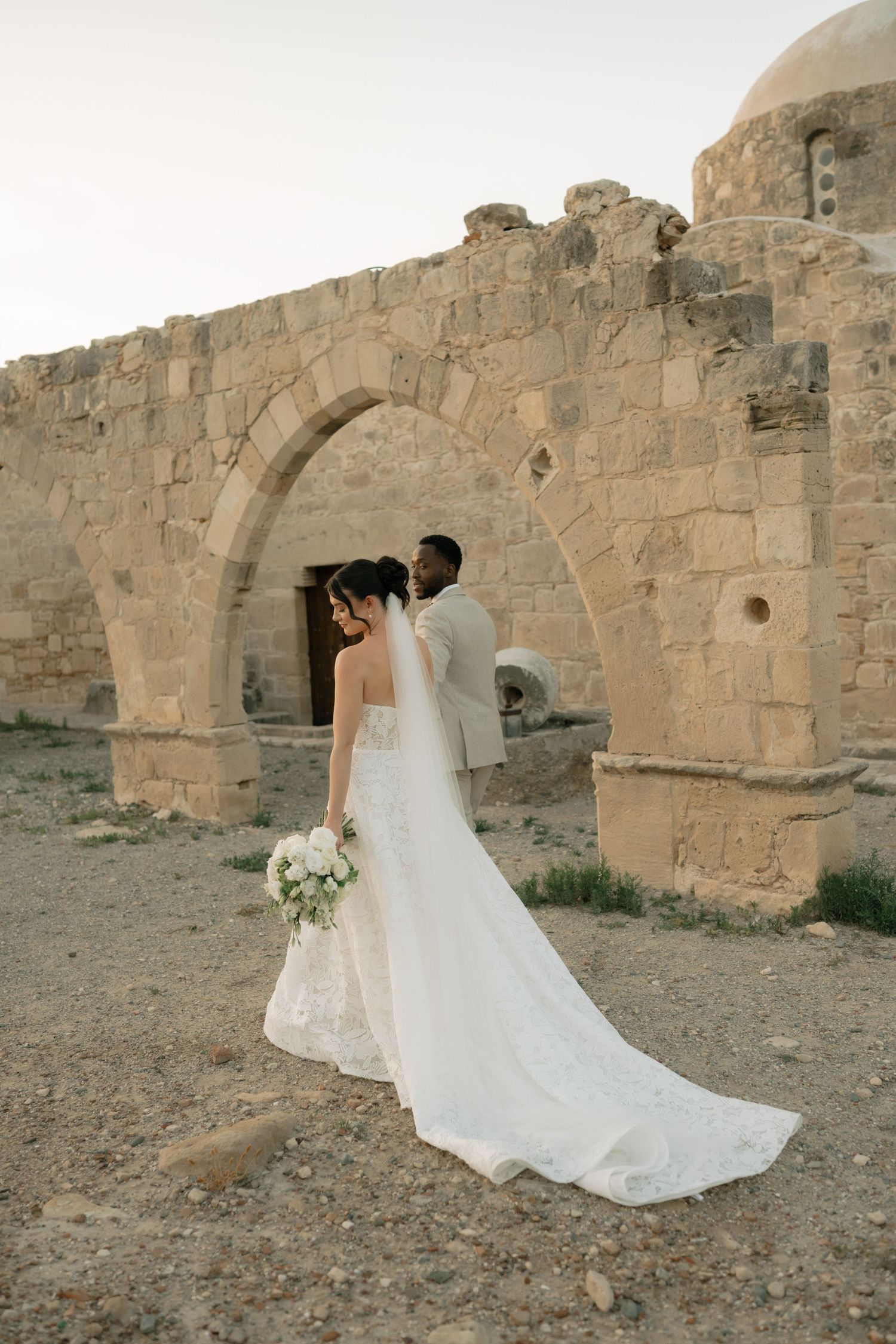 Bride and groom pose at ancient stone arch ruins in desert landscape, bride wearing elegant white gown with long train.