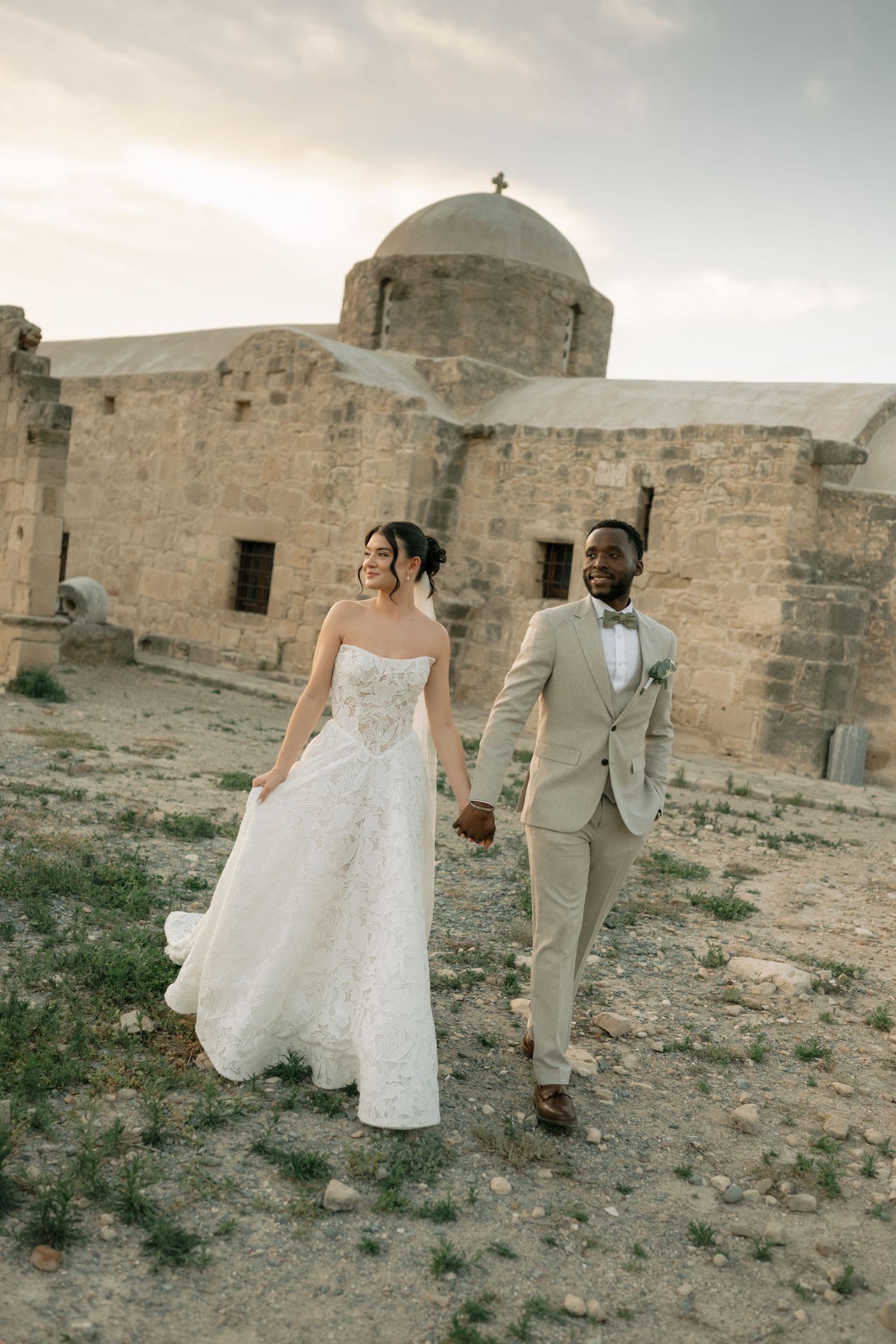 Interracial couple in wedding attire holding hands walking near ancient stone church at sunset.
