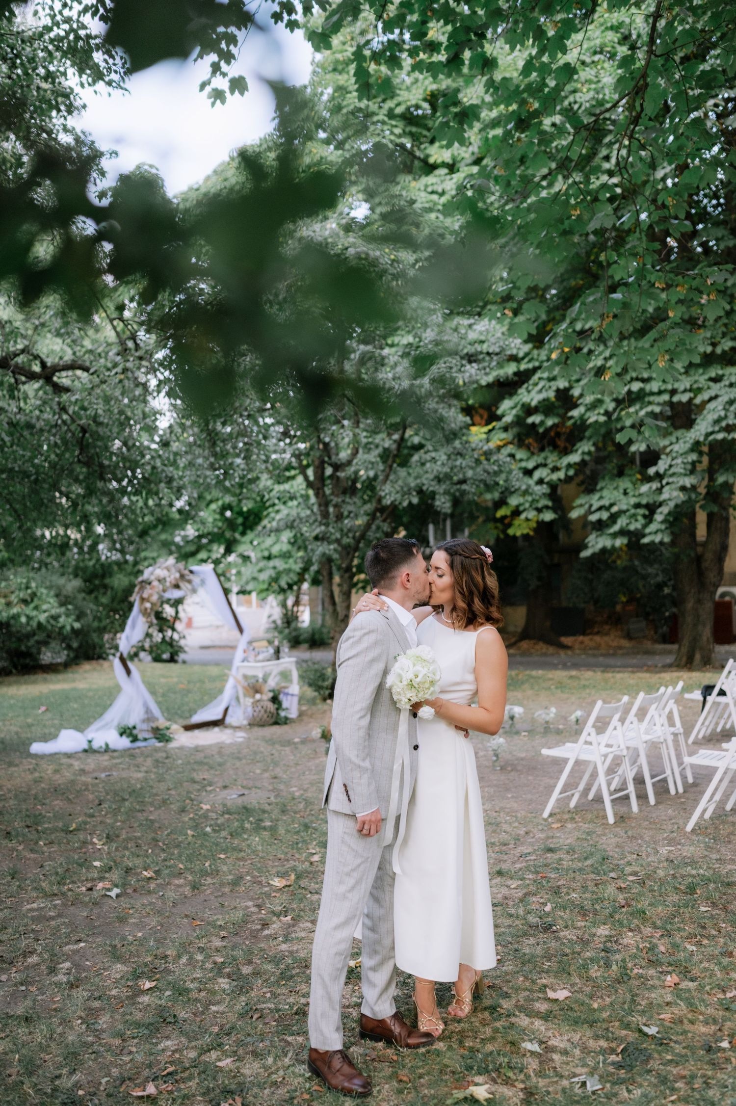 A romantic outdoor wedding ceremony setup in a wooded area with white chairs and natural greenery surroundings.