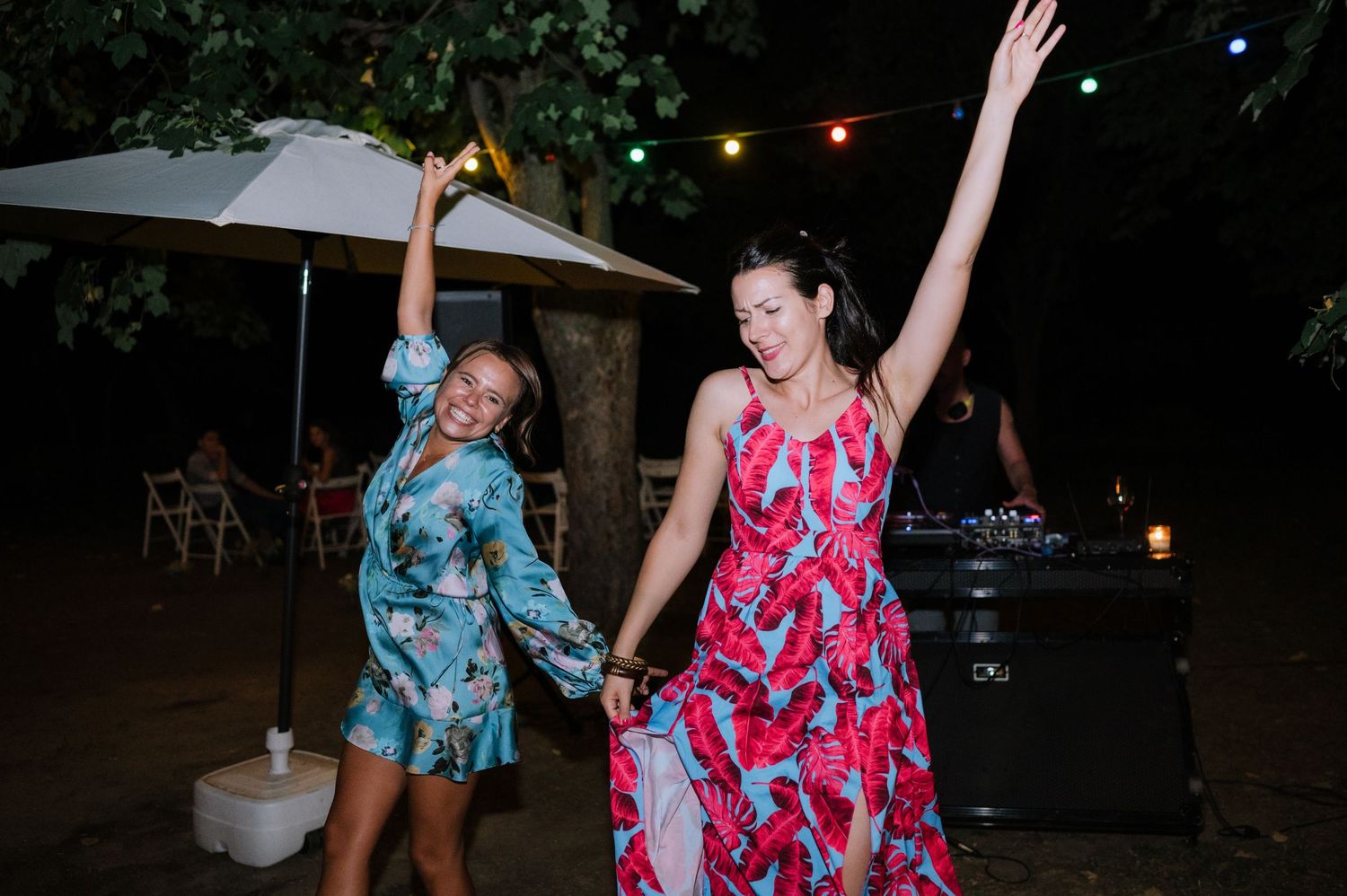 Two people in colorful floral dresses dance together at an outdoor evening event with string lights overhead.