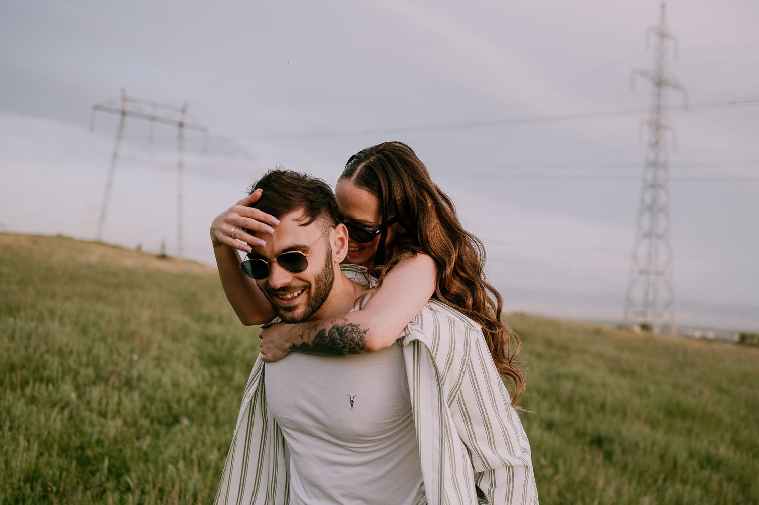 A couple embraces in a grassy field with power lines and a cloudy sky in the background.