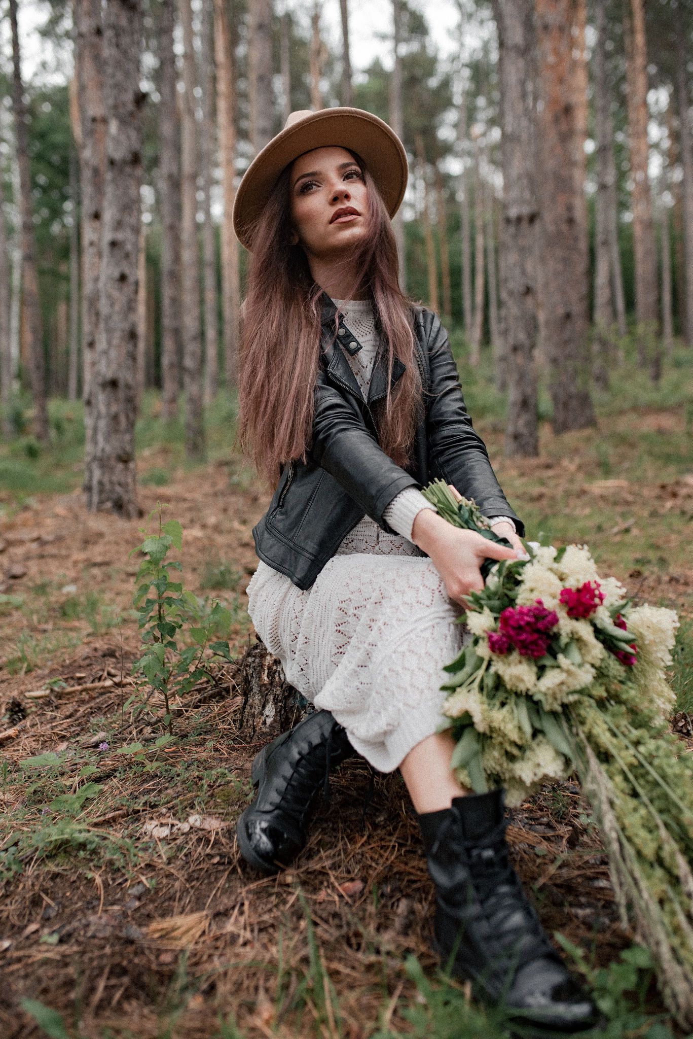 Stylish figure in white dress and leather jacket sits in forest holding burgundy and cream floral bouquet.