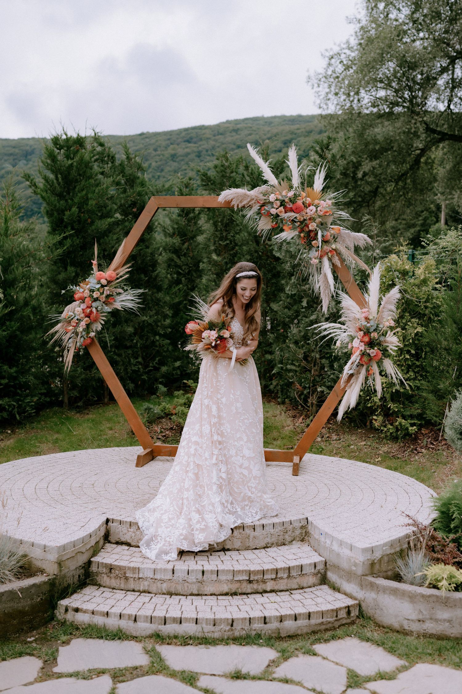 A rustic outdoor wedding ceremony setup with a hexagonal wooden arch decorated with dried flowers and pampas grass on stone steps.