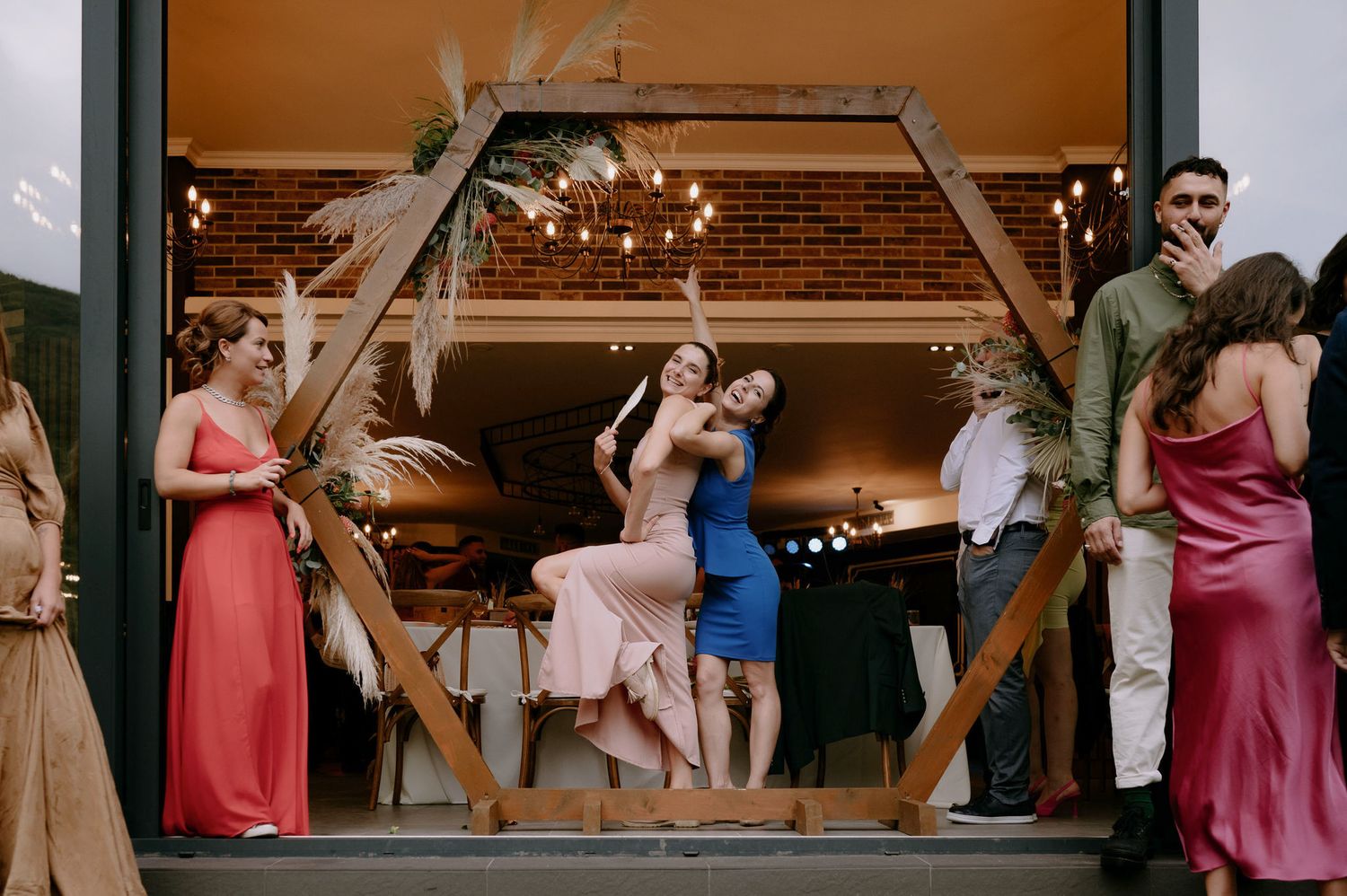 A group of people dance and celebrate at a wedding reception in a rustic venue with exposed wooden beams.