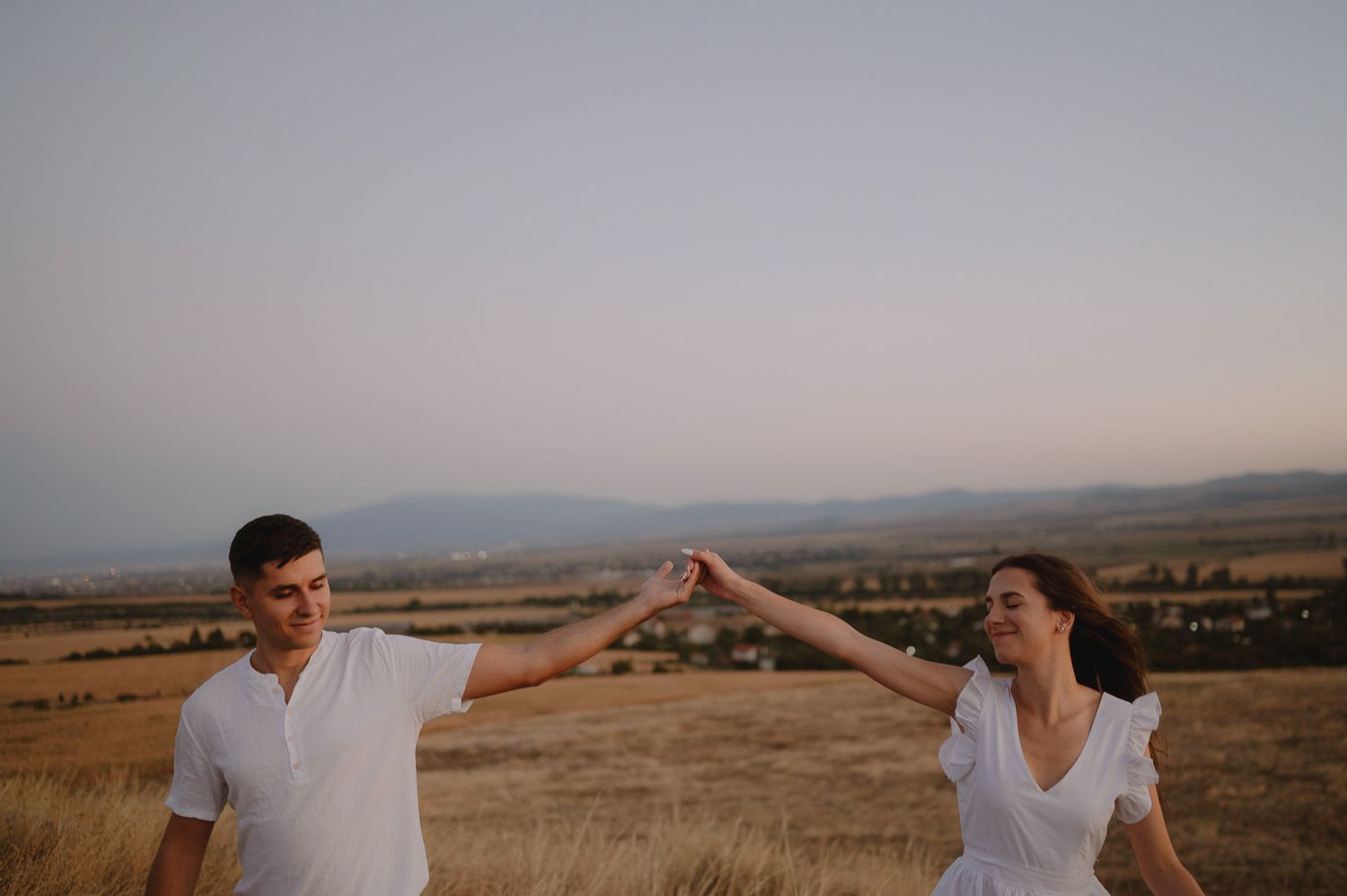 A couple in white outfits holds hands in a golden field at sunset with mountains in the distance.
