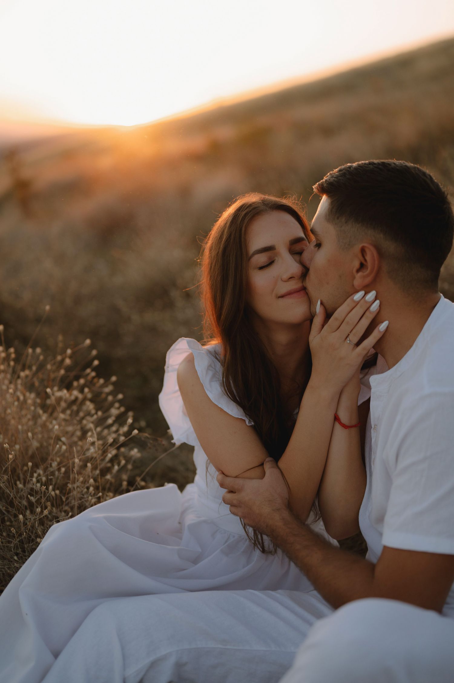 Romantic couple in white outfits share an intimate moment at sunset in a golden field.
