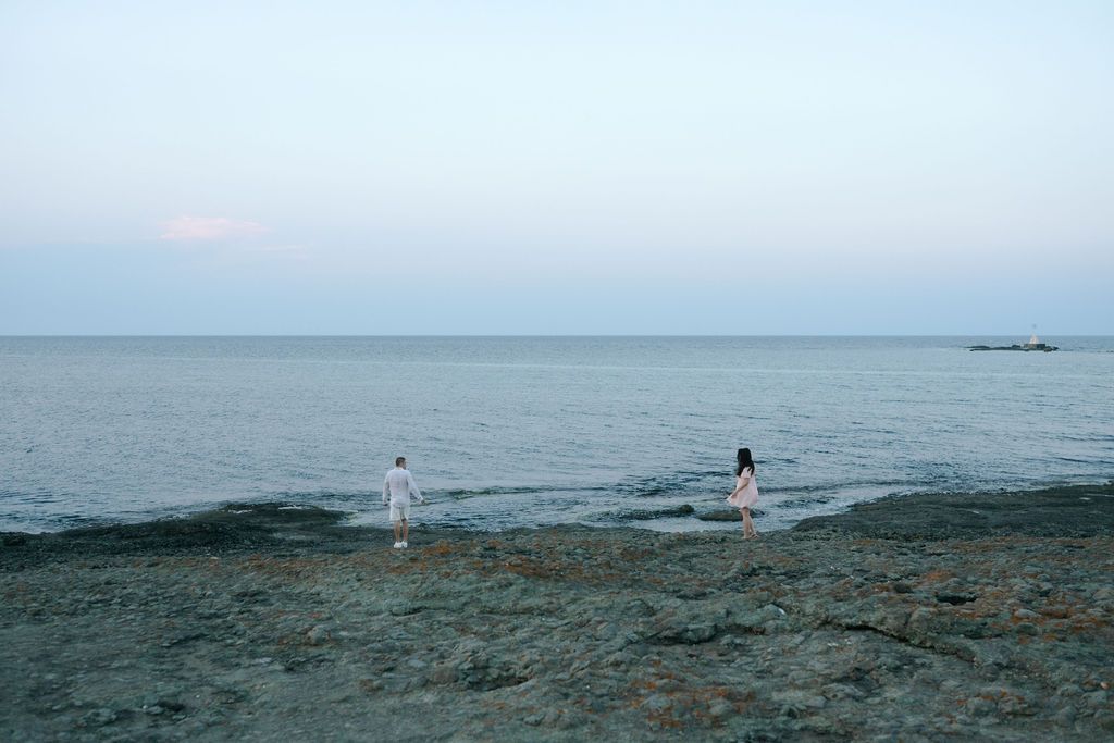 Two people stand at the edge of a rocky beach looking out over the calm ocean at dusk.
