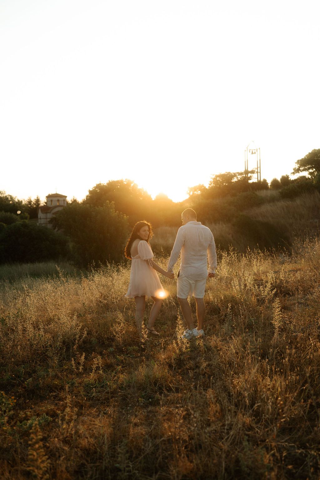 A couple walks hand in hand through a golden field at sunset, backlit by the warm glowing sun.