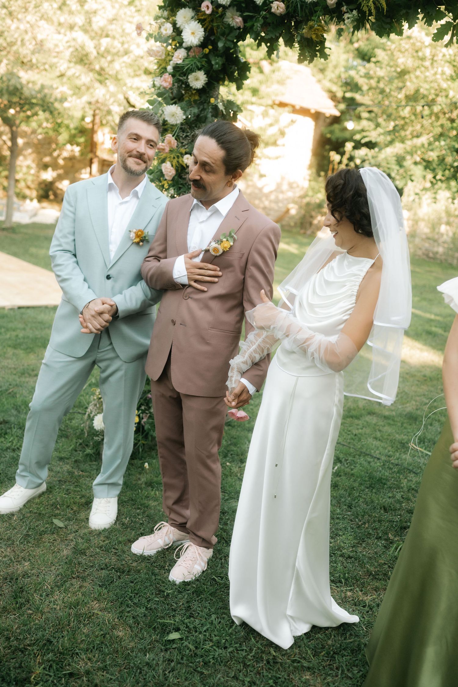 Wedding party members in coordinated mint green and brown suits share joyful moments on a grassy lawn.