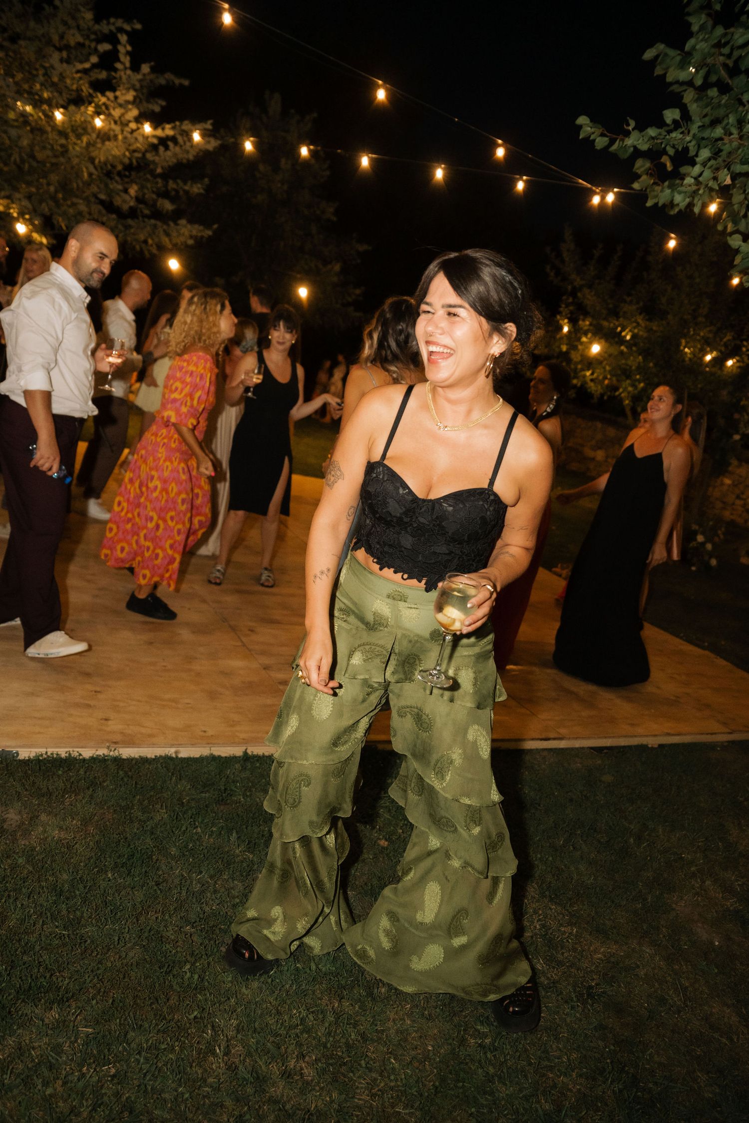 A person in a green dress dances joyfully under string lights at an outdoor evening party.