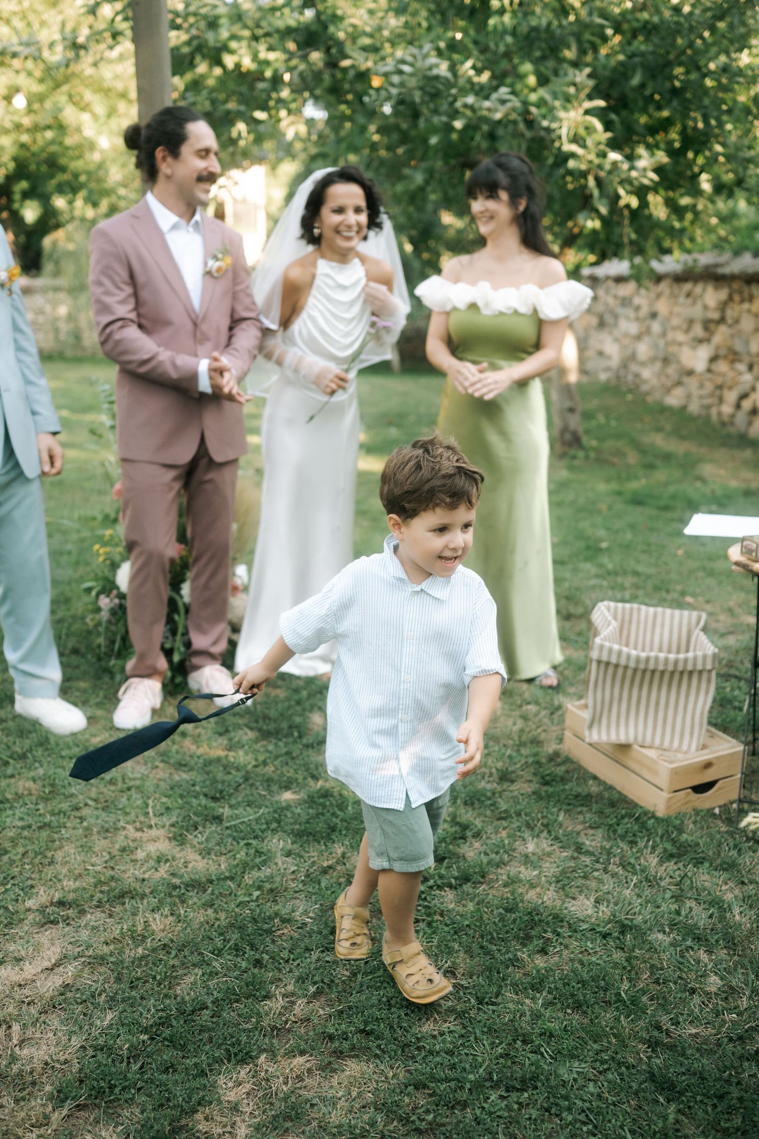 A young child in a white shirt and shorts stands on grass while wedding attendees in formal attire gather behind.