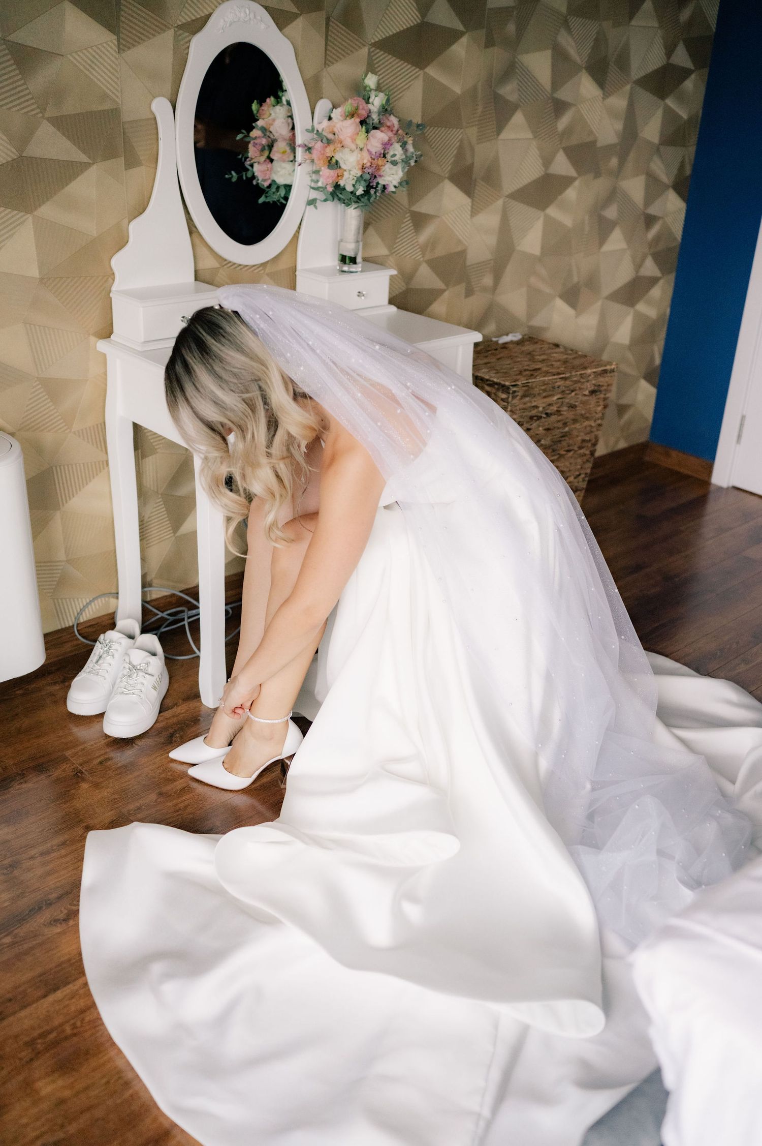 A bride in a flowing white wedding dress sits at a vanity putting on her shoes before the ceremony.