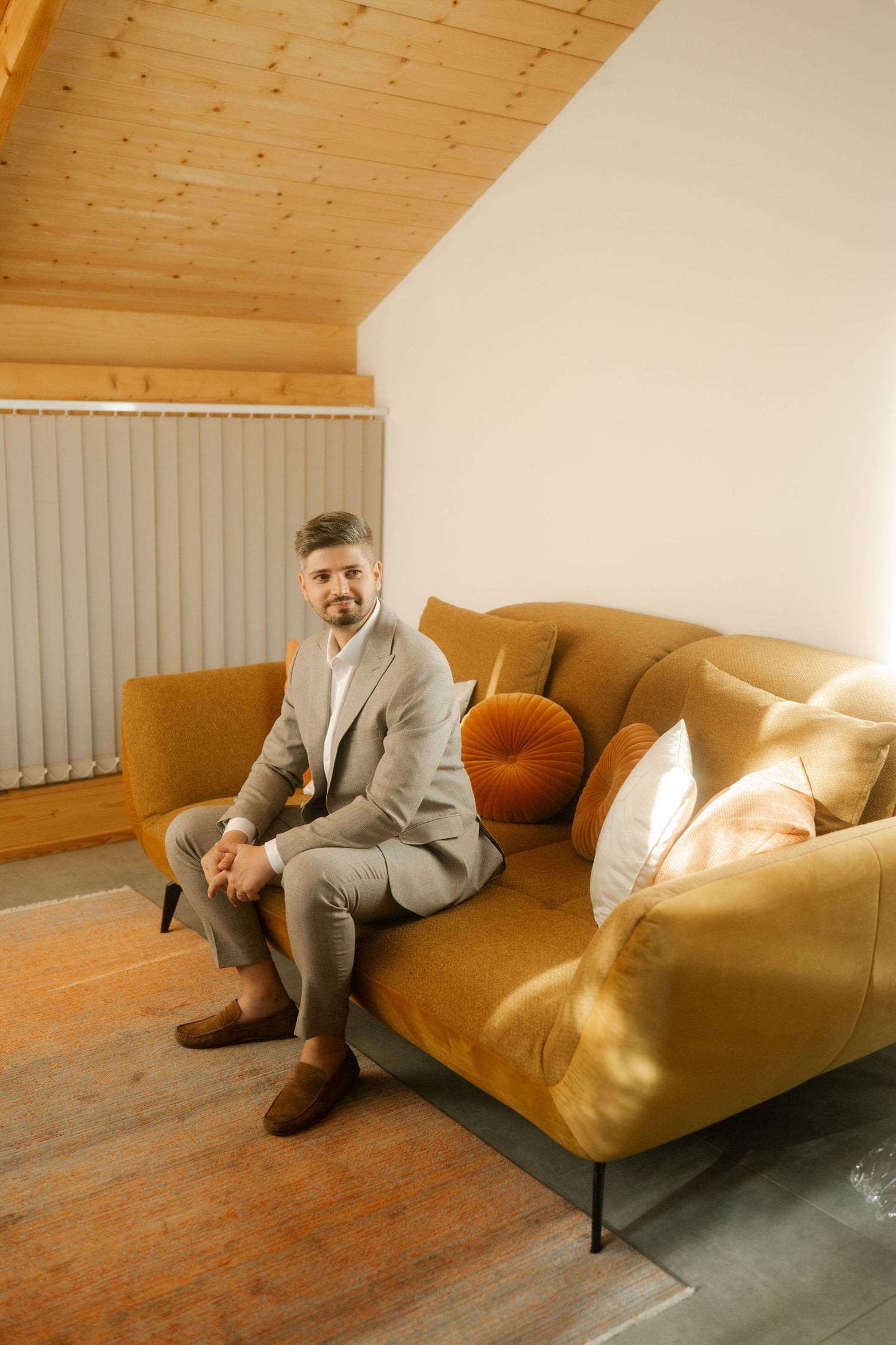 A person in a gray suit lounges on a tan leather sofa in a room with wooden floors and ceiling.