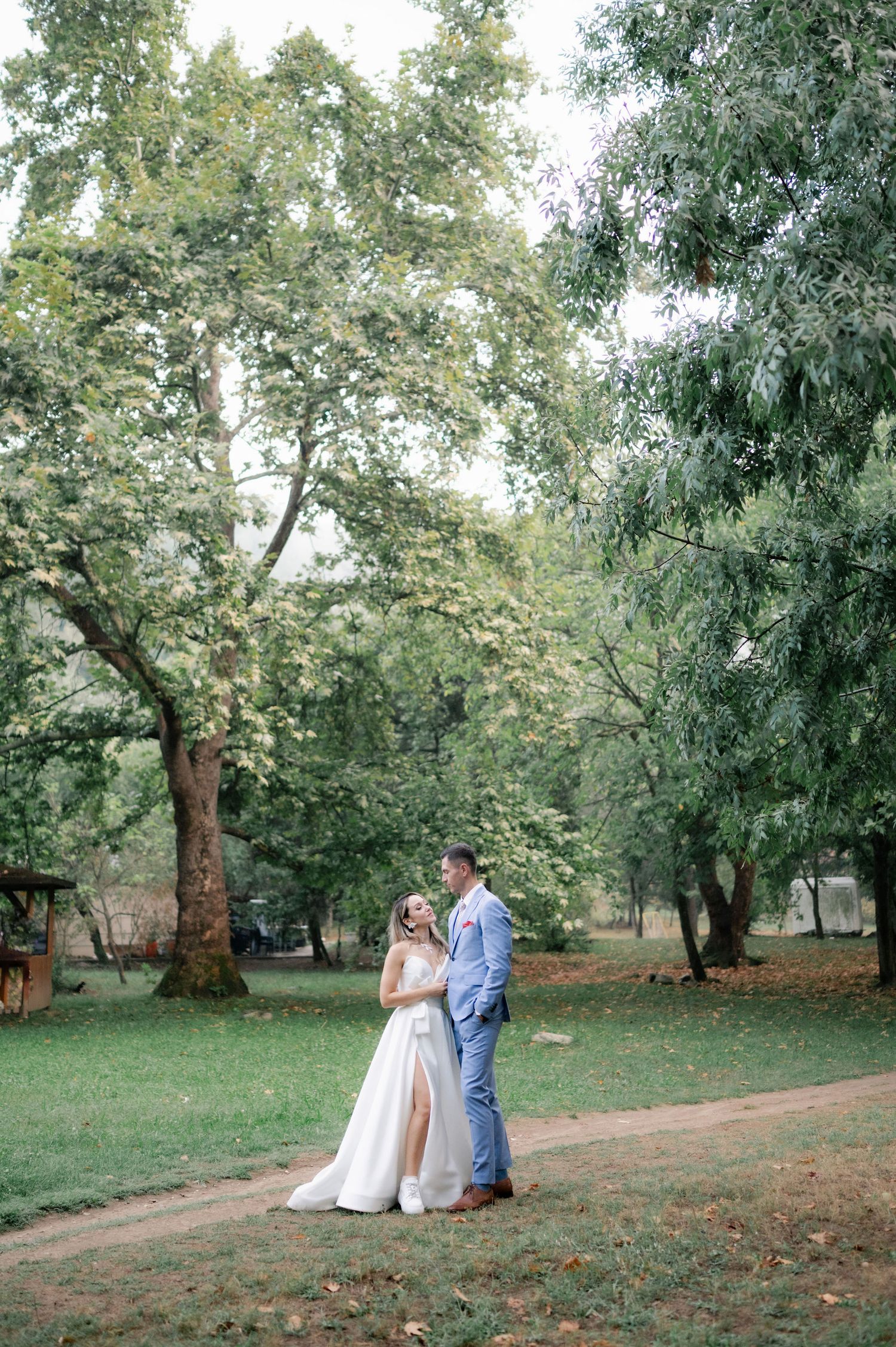 A couple shares tender moments together in a serene park setting with lush green trees in the background.