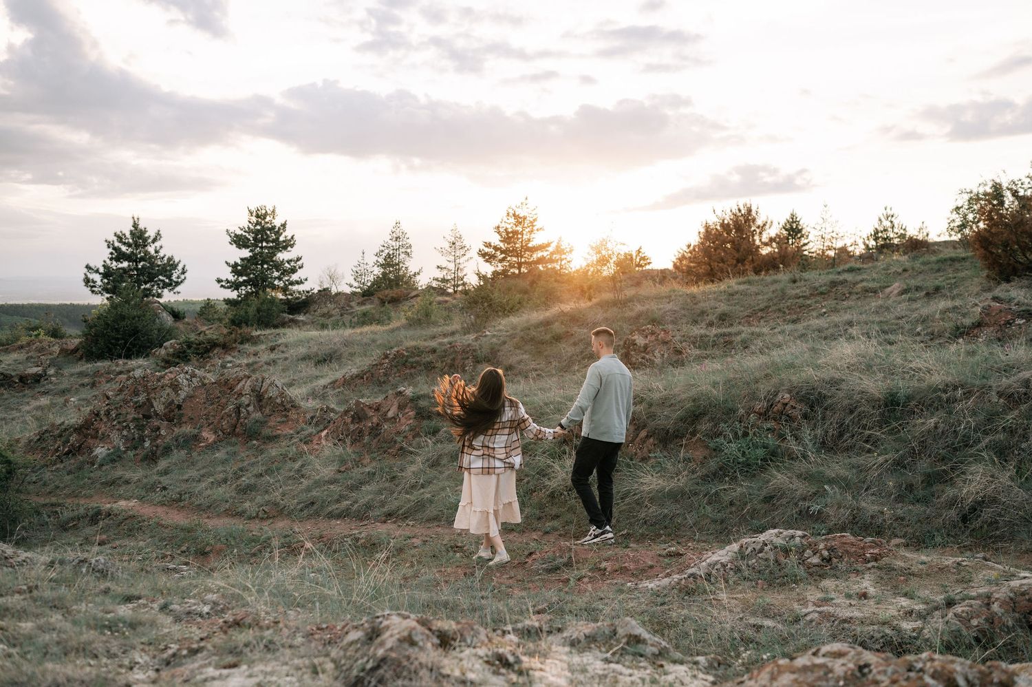 Couple walks along scenic hillside path at sunset while holding hands as golden light illuminates the landscape.