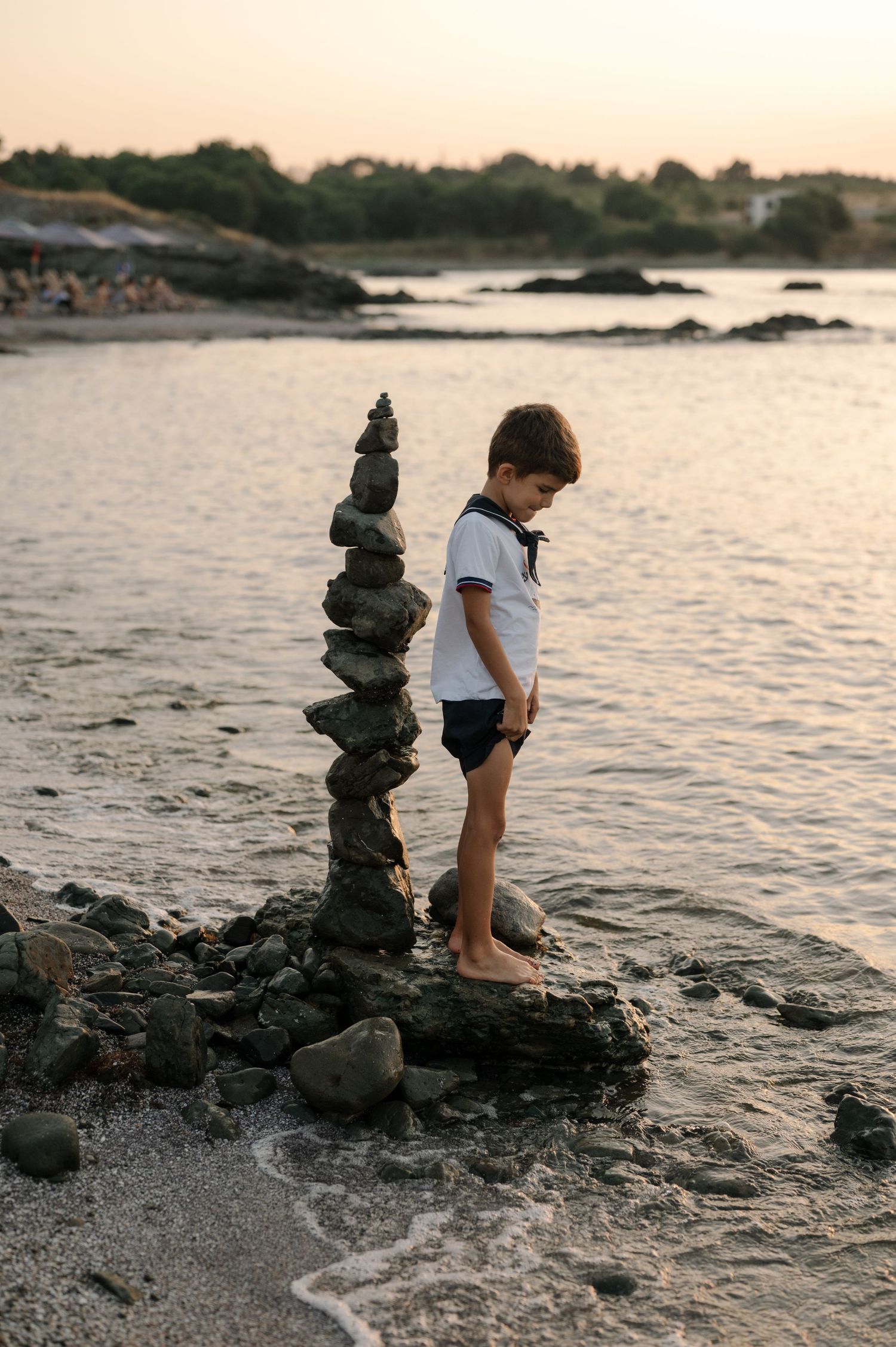 A small figure stands beside a tall stack of balanced rocks on a beach at sunset with waves lapping at the shore.