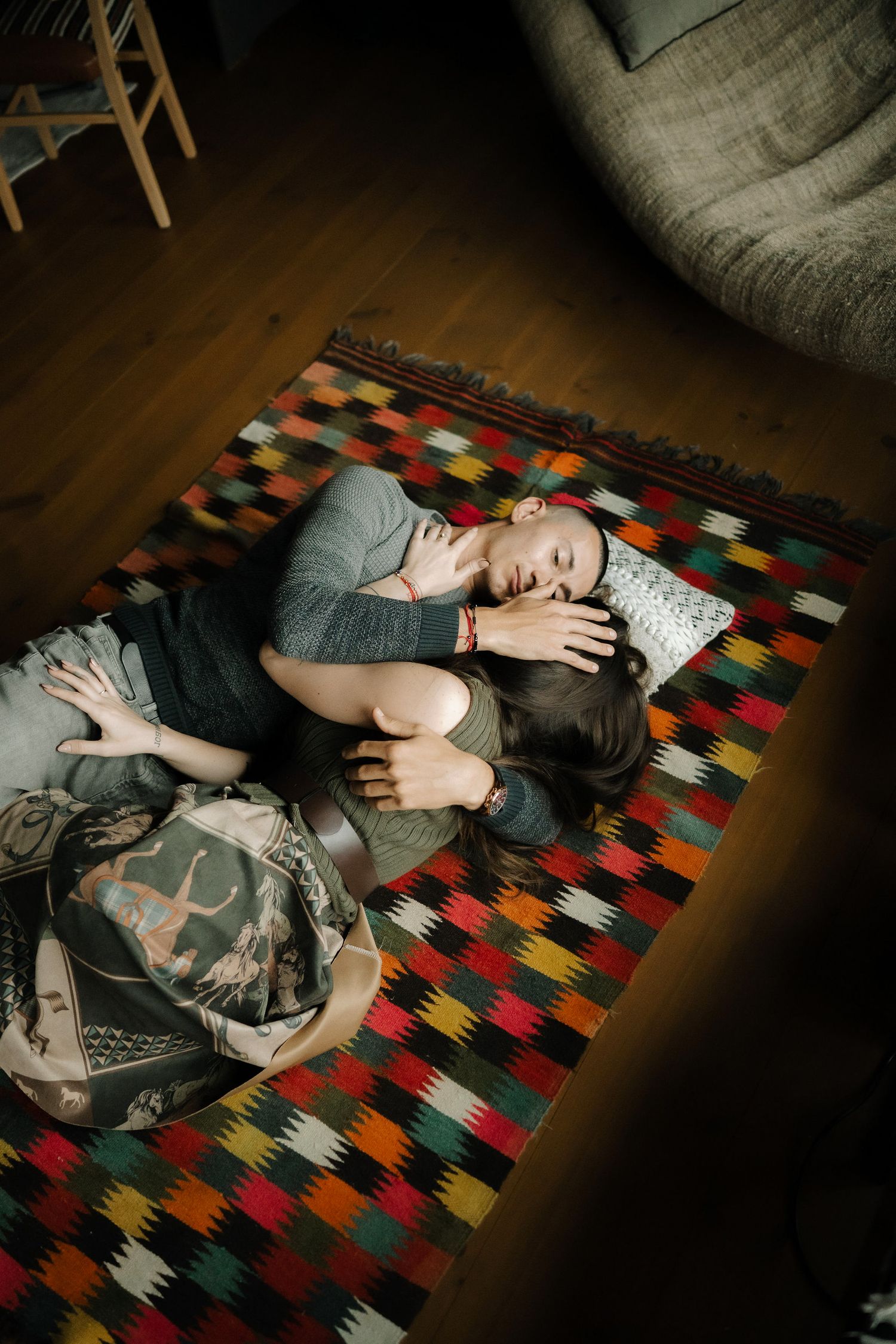 A sleeping figure curled up on a colorful geometric patterned rug with a gray pillow and dark wooden floor.