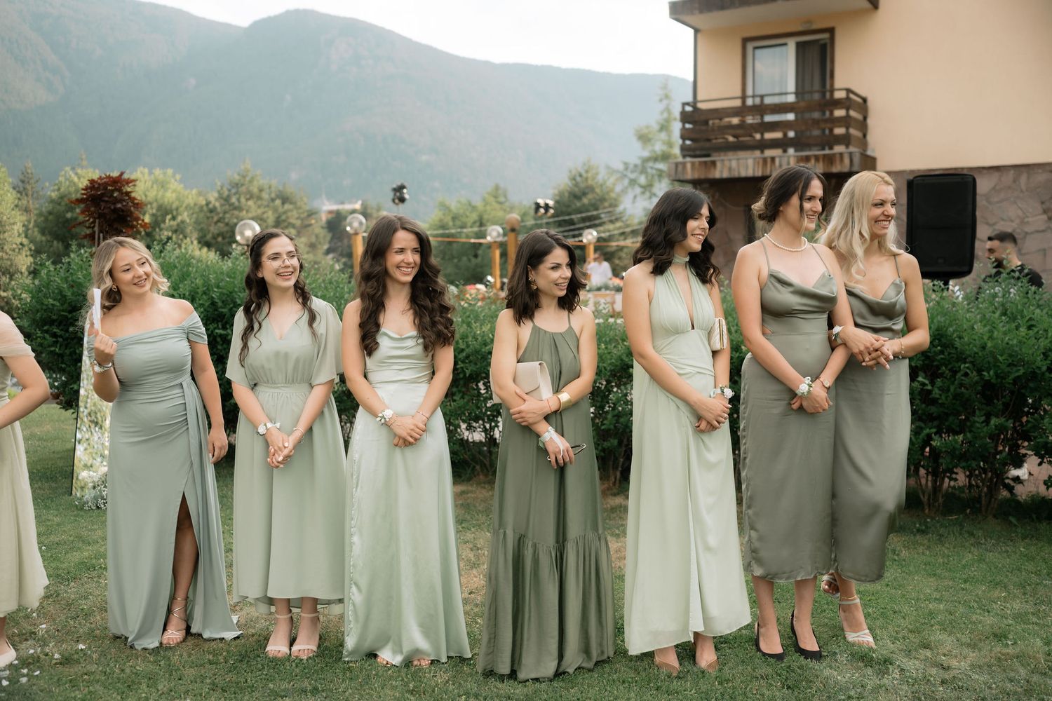 A row of people in sage green and cream colored dresses stand together outdoors with mountains in the background.