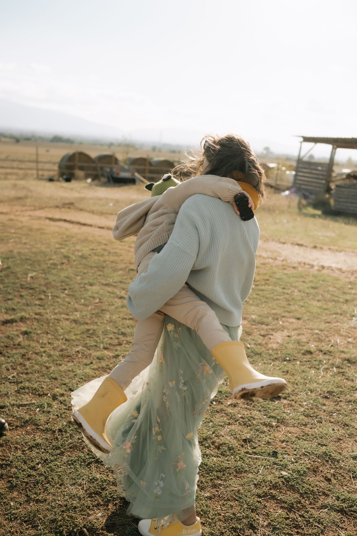 A person in a light-colored coat and dress spins joyfully in a sunlit field at golden hour.