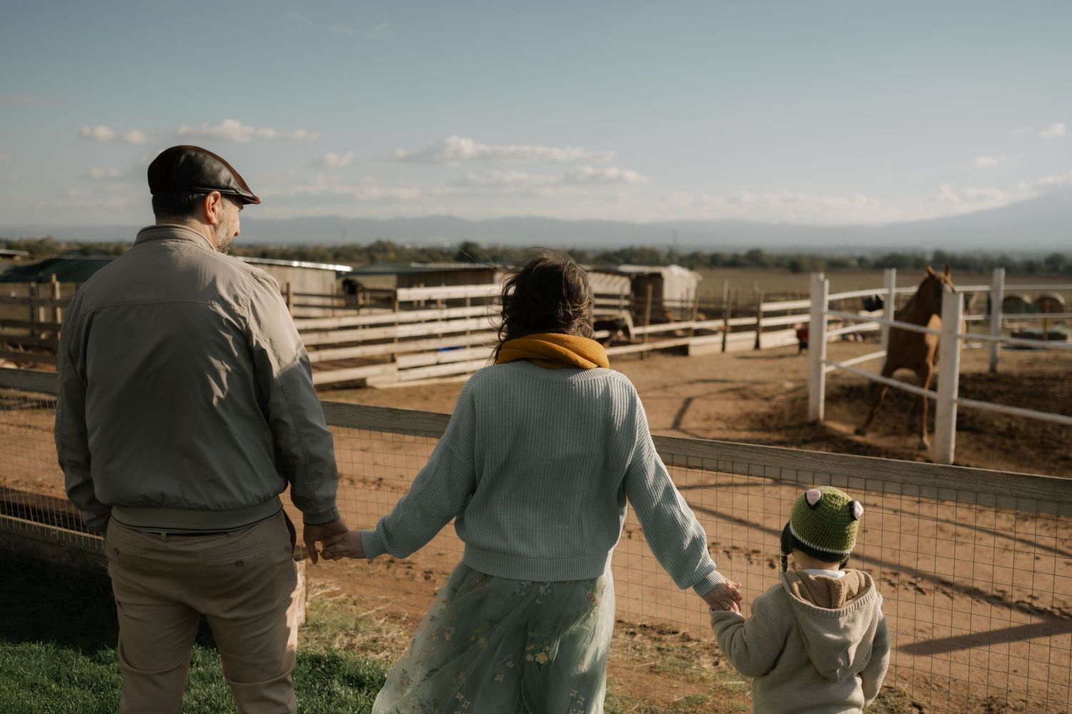 Two people stand at a wooden fence overlooking a scenic rural landscape on a sunny afternoon.