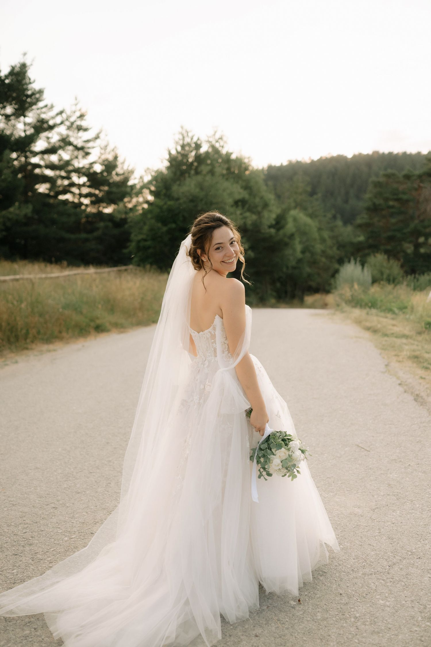 A bride in a flowing white gown twirls gracefully on a sunlit path surrounded by evergreen trees.