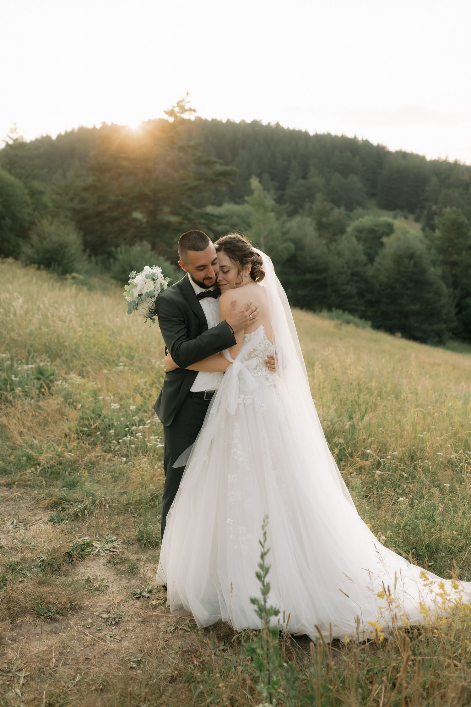 A romantic wedding couple embraces in a golden sunlit meadow at sunset while wearing formal attire.