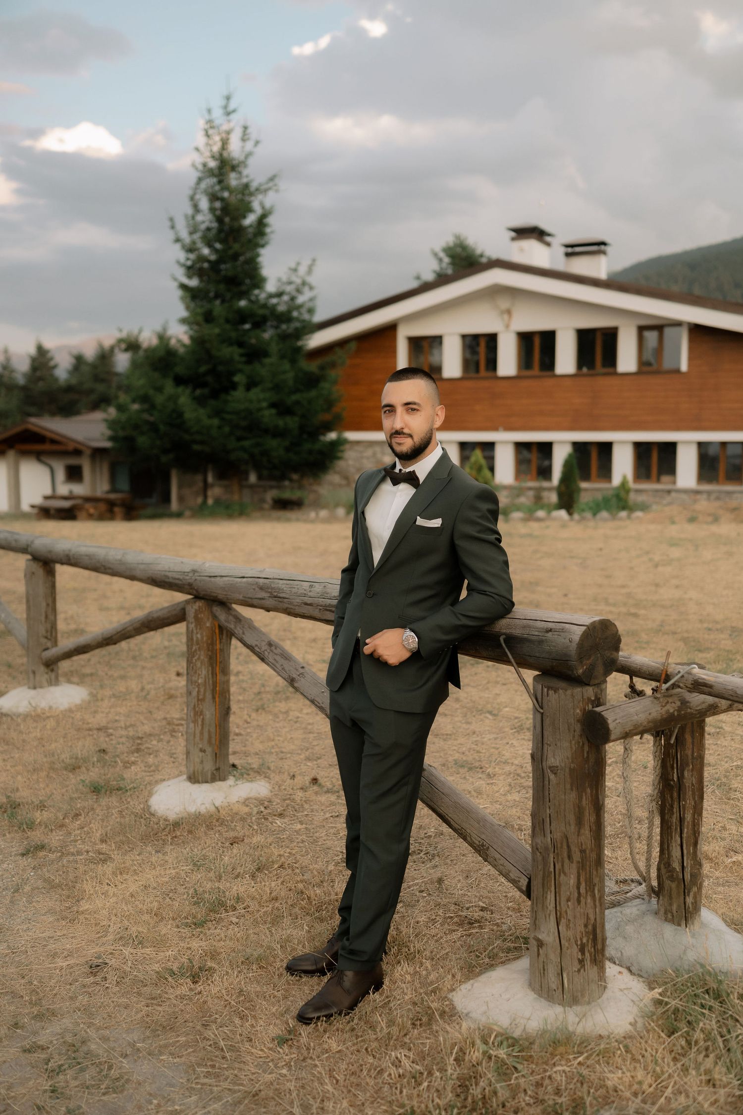 A groom in a black suit poses casually against a rustic wooden fence on a rural property with buildings in the background.