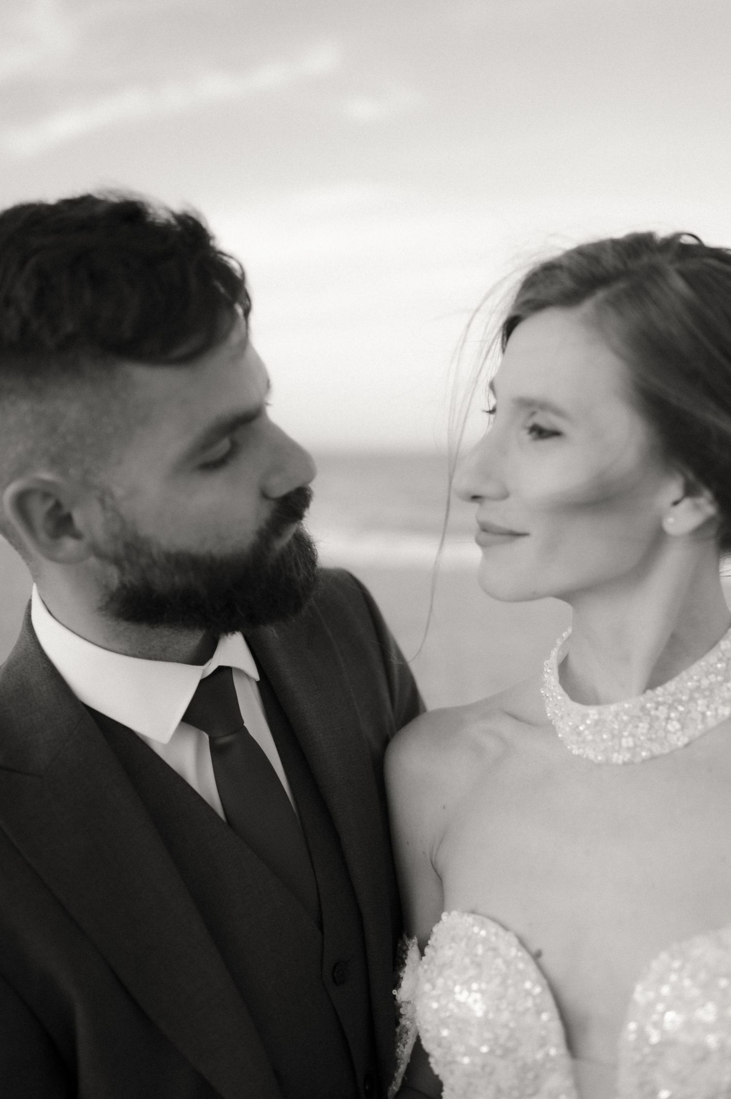 Black and white wedding portrait of a bearded groom in a suit and bride in a beaded gown gazing at each other on a beach.