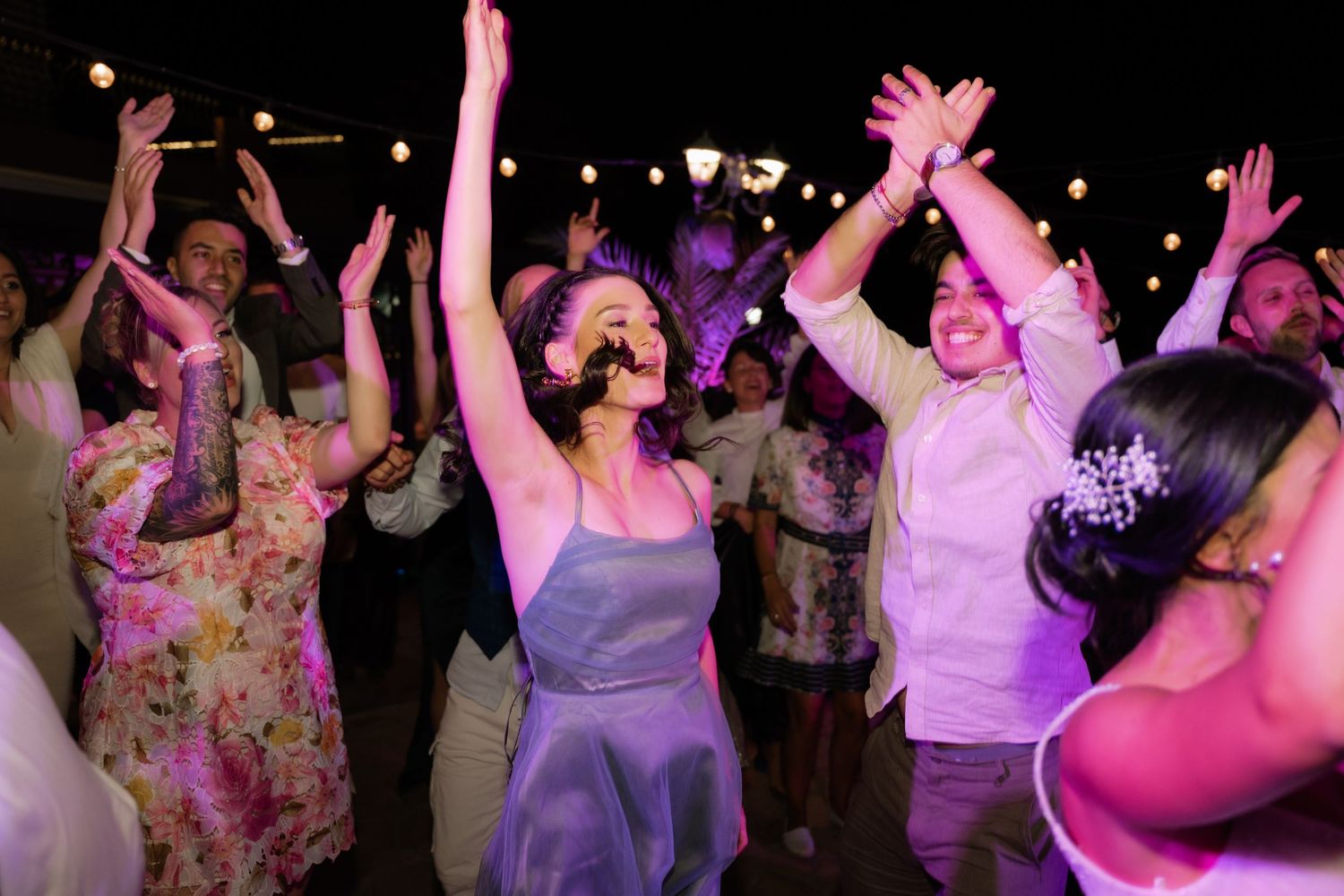 A crowd of party guests dance with raised arms under pink lighting at a nighttime celebration.