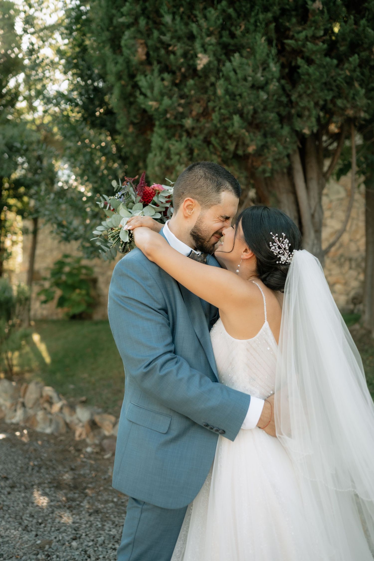A romantic wedding couple embraces in an outdoor garden setting with lush greenery in the background.