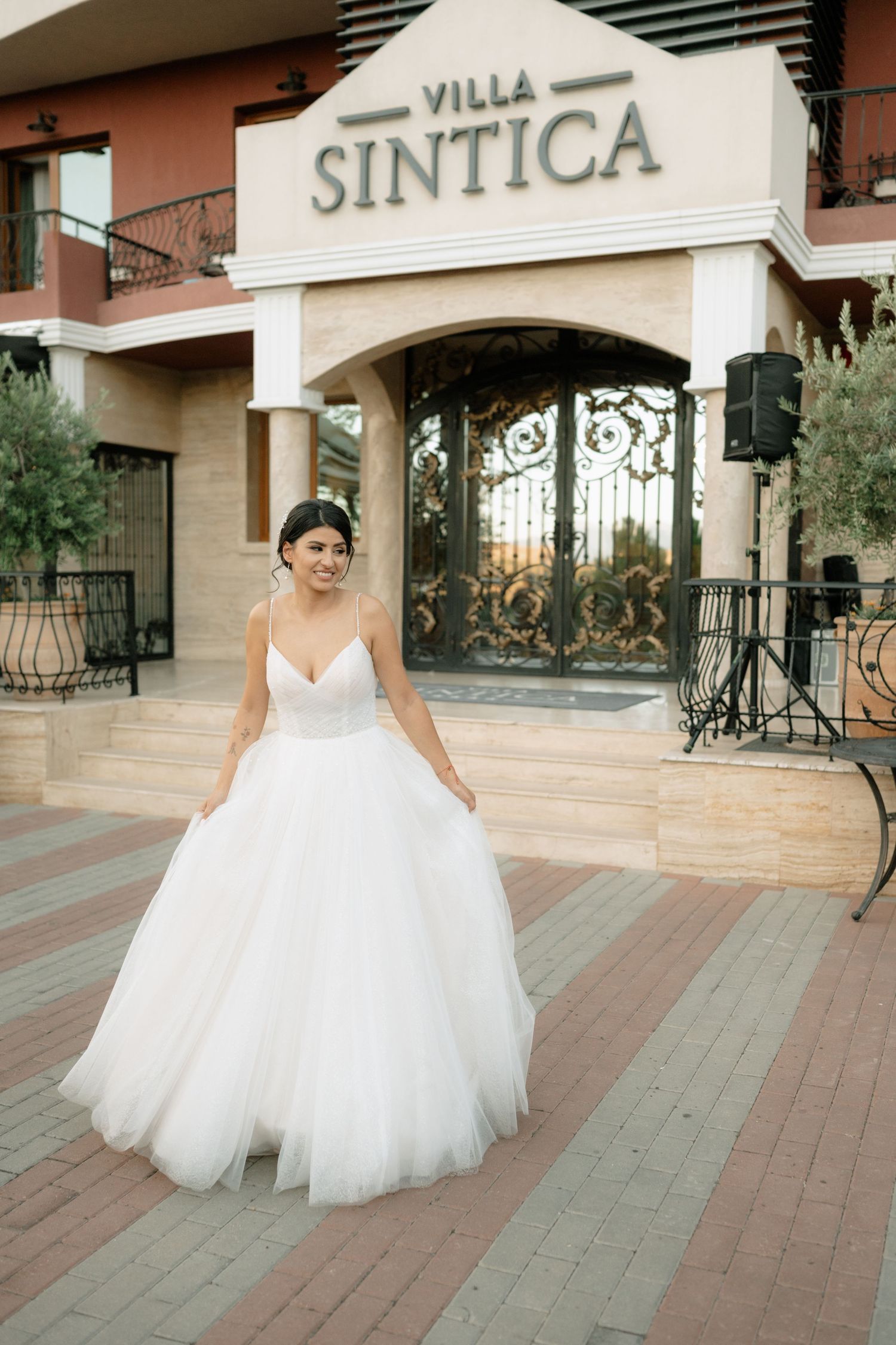 A bride in a flowing white tulle ballgown poses elegantly outside a villa entrance with ornate iron gates.