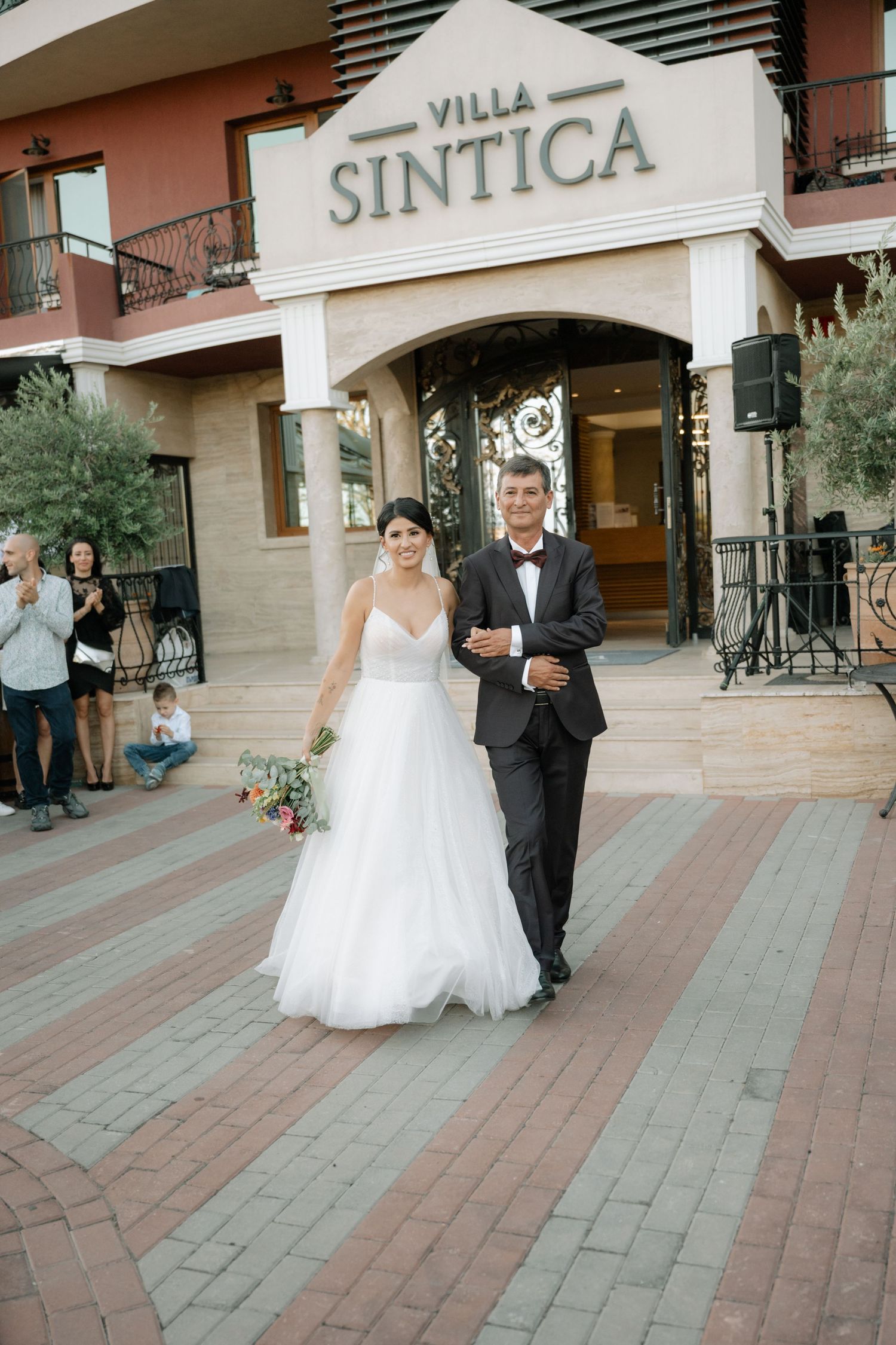 A wedding couple exits Villa Sintica venue on a brick paved walkway with guests watching in the background.