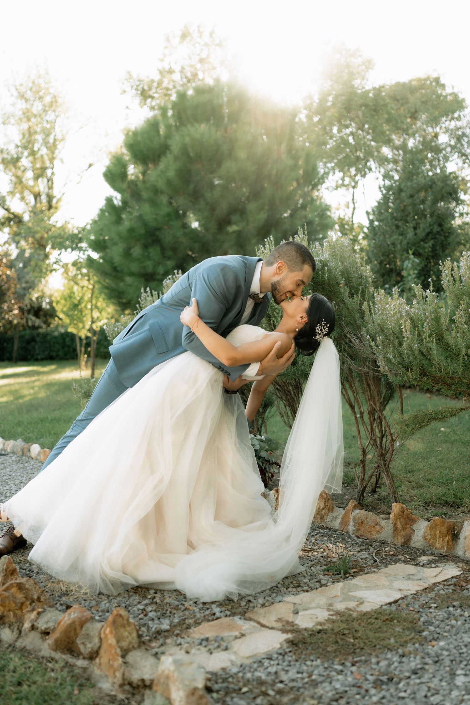 A romantic wedding couple shares a kiss in a garden setting with sunlight filtering through trees in the background.