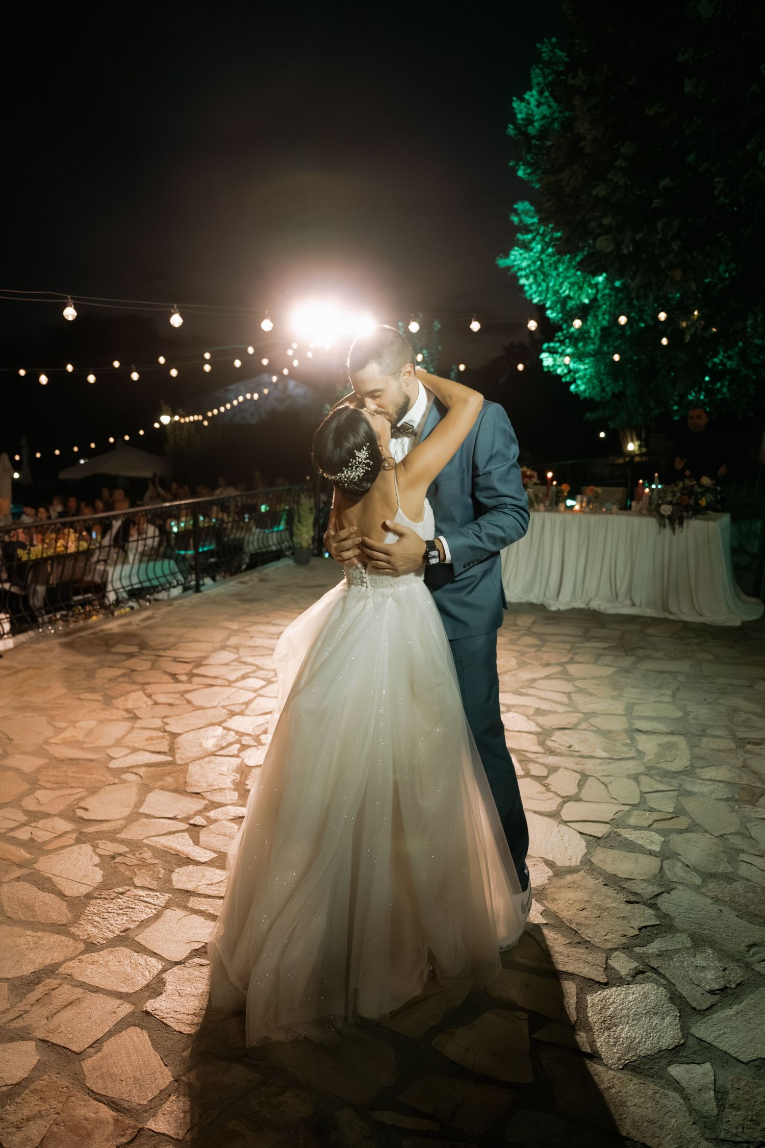 A romantic first dance at an outdoor evening wedding reception with string lights and stone patio flooring.