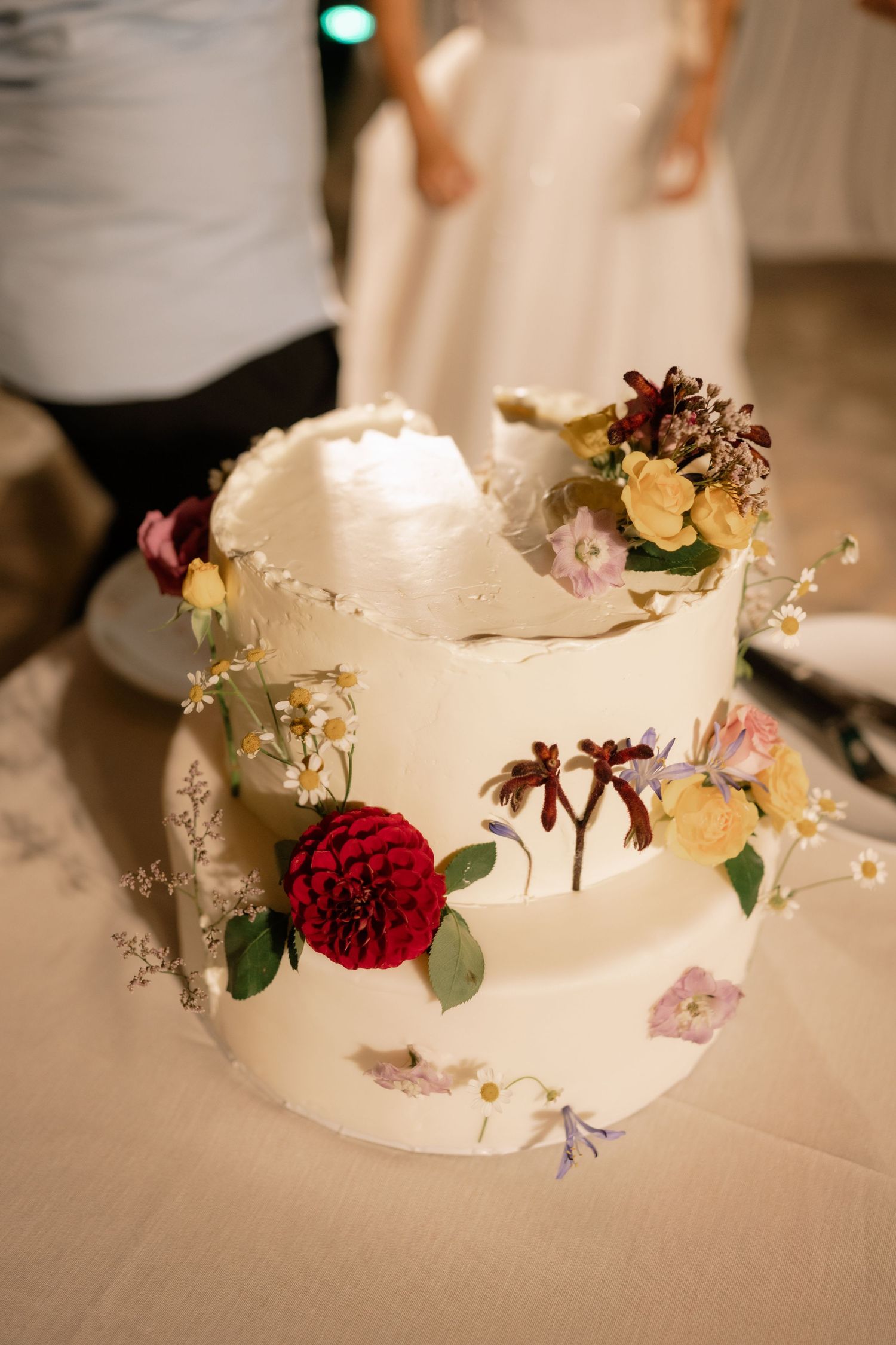 A white tiered wedding cake decorated with fresh flowers including a red dahlia, yellow blooms and delicate baby's breath.
