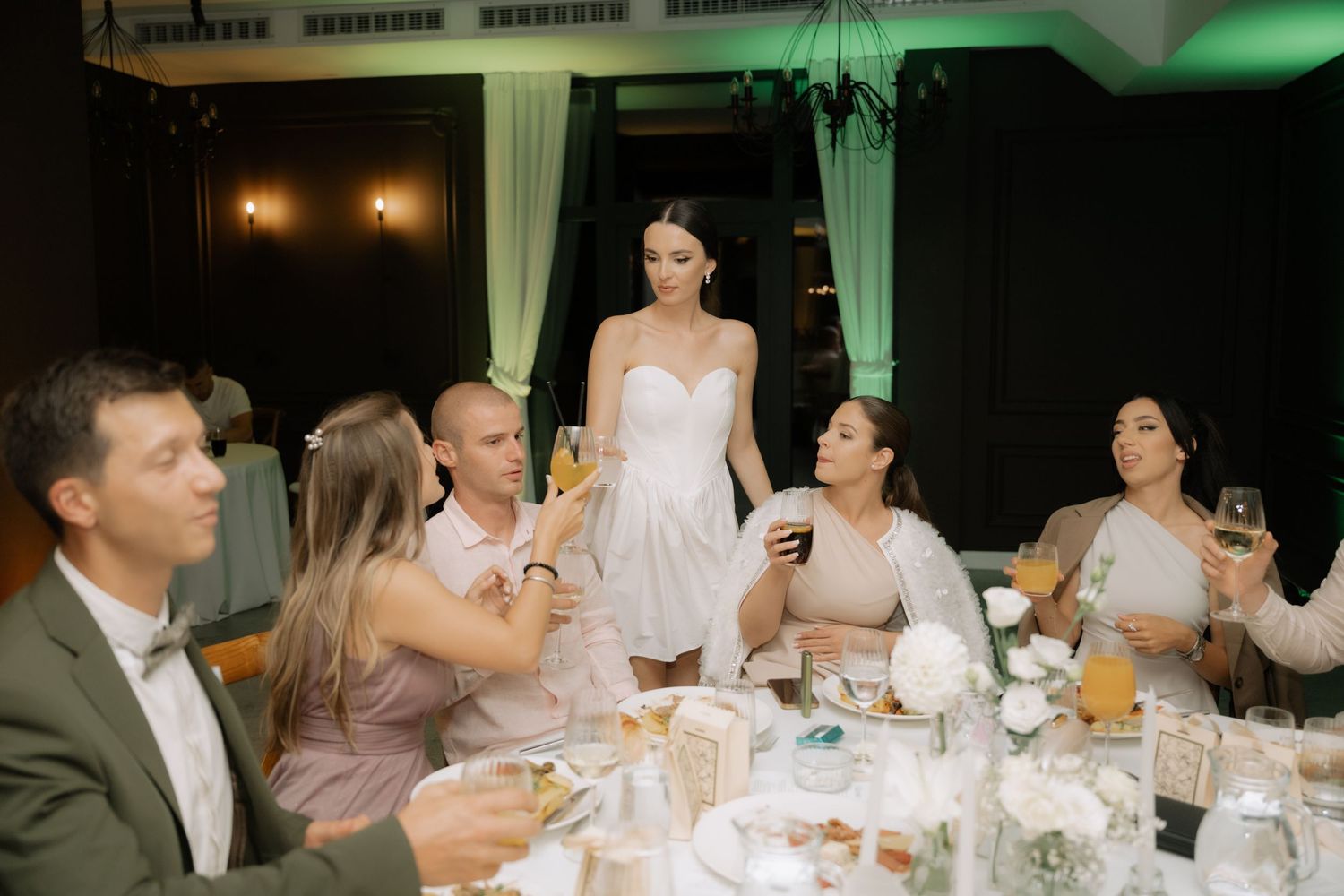 Wedding guests seated at a formal dinner table in a reception hall with green accent lighting and white decor.