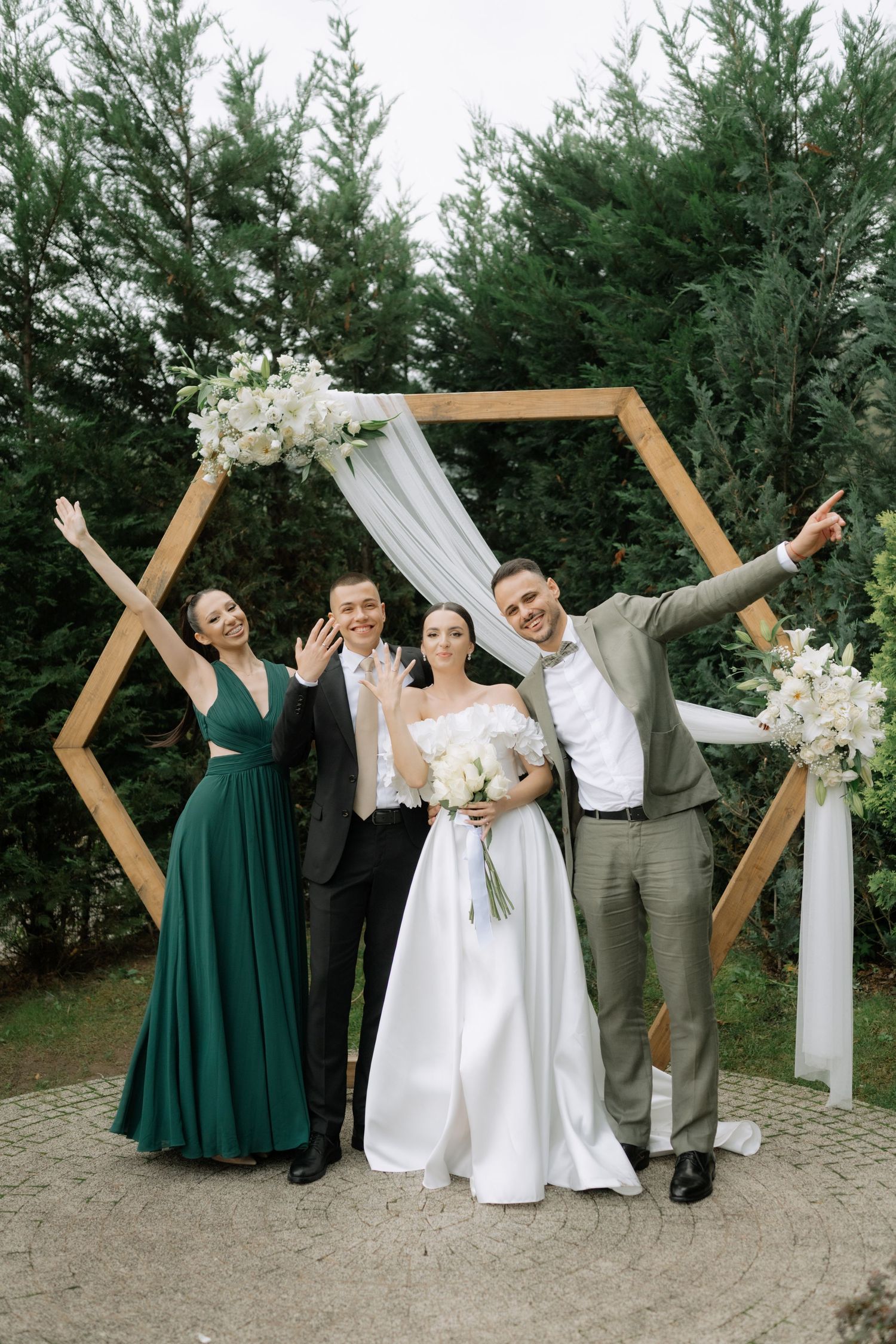Joyful wedding party celebrating under a modern hexagonal arch with white floral arrangements and green foliage.