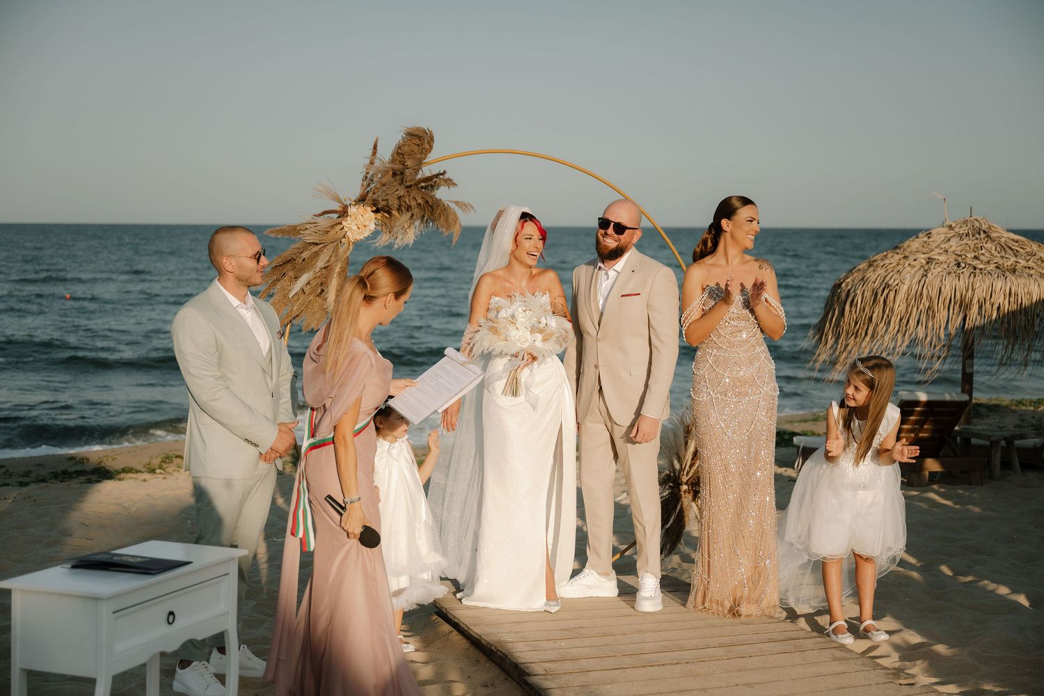 A beachside wedding celebration with people in white and beige attire walking along the sandy shore at sunset.